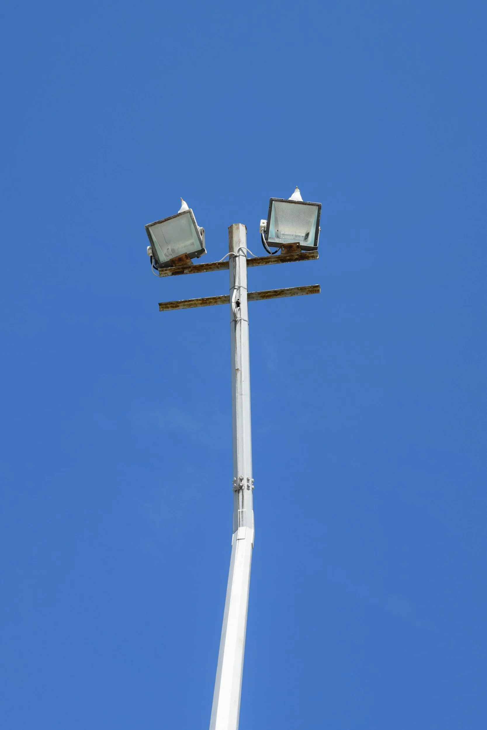 A tall pole with two floodlights attached at the top, against a clear blue sky. The floodlights are slightly rusted with some snow on top.