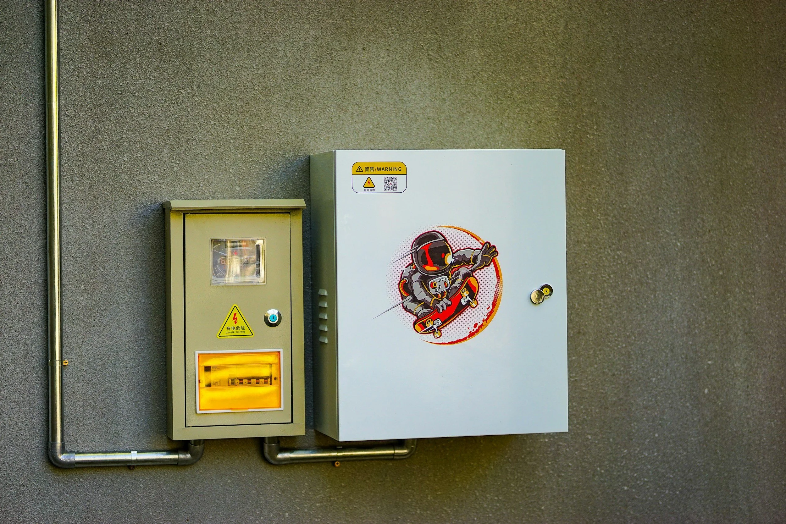 Electrical boxes mounted on a wall with warning labels, one has a cartoon astronaut skateboarding sticker.