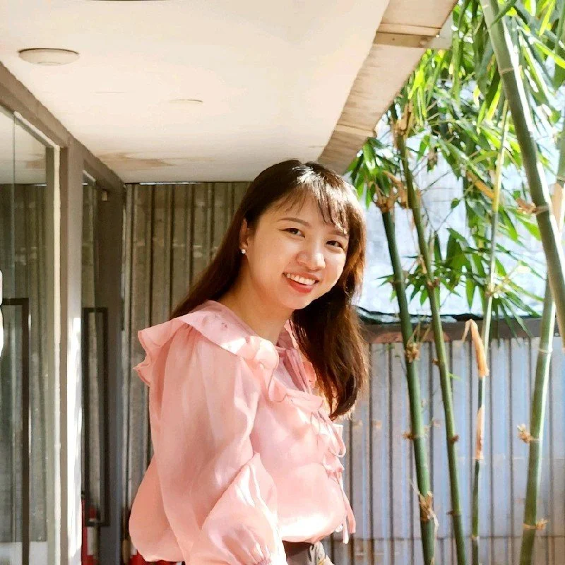 A woman with shoulder-length black hair and a bright smile, wearing a blue top, standing among green leafy plants indoors.