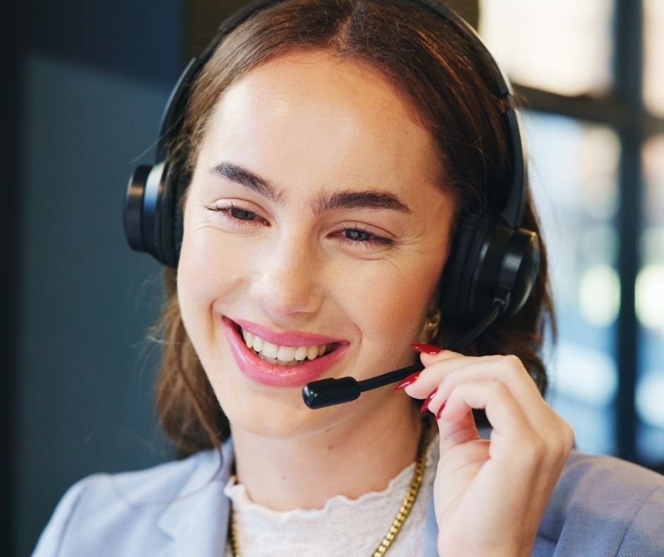 A man with brown curly hair, a beard, and glasses, wearing a black sweater, looks to the side with a slight smile. Background includes a white wall and a blurred green plant.