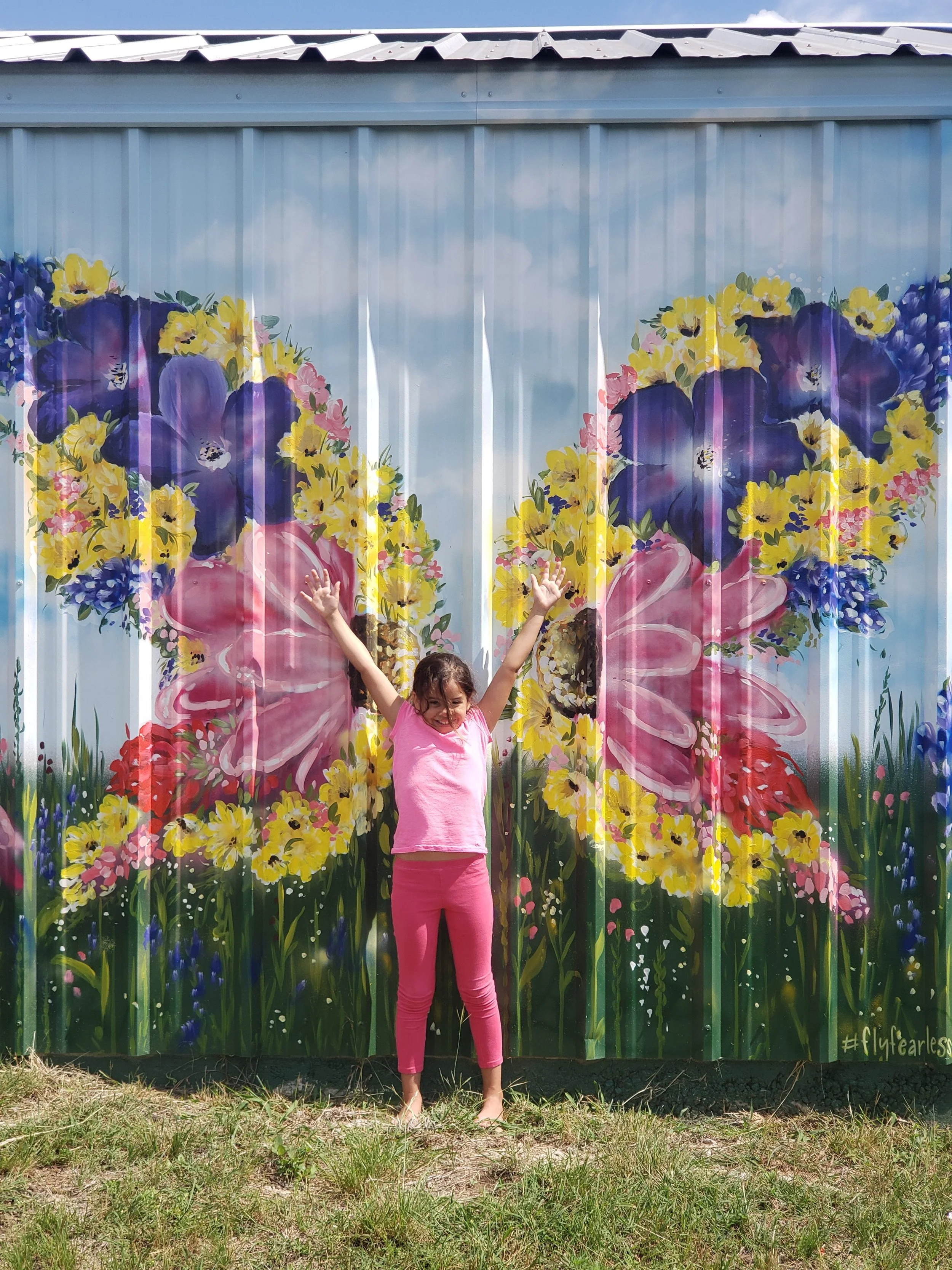 Girl in pink clothes with arms raised standing in front of a colorful mural of large purple, pink, yellow, and red flowers painted on a metal fence.