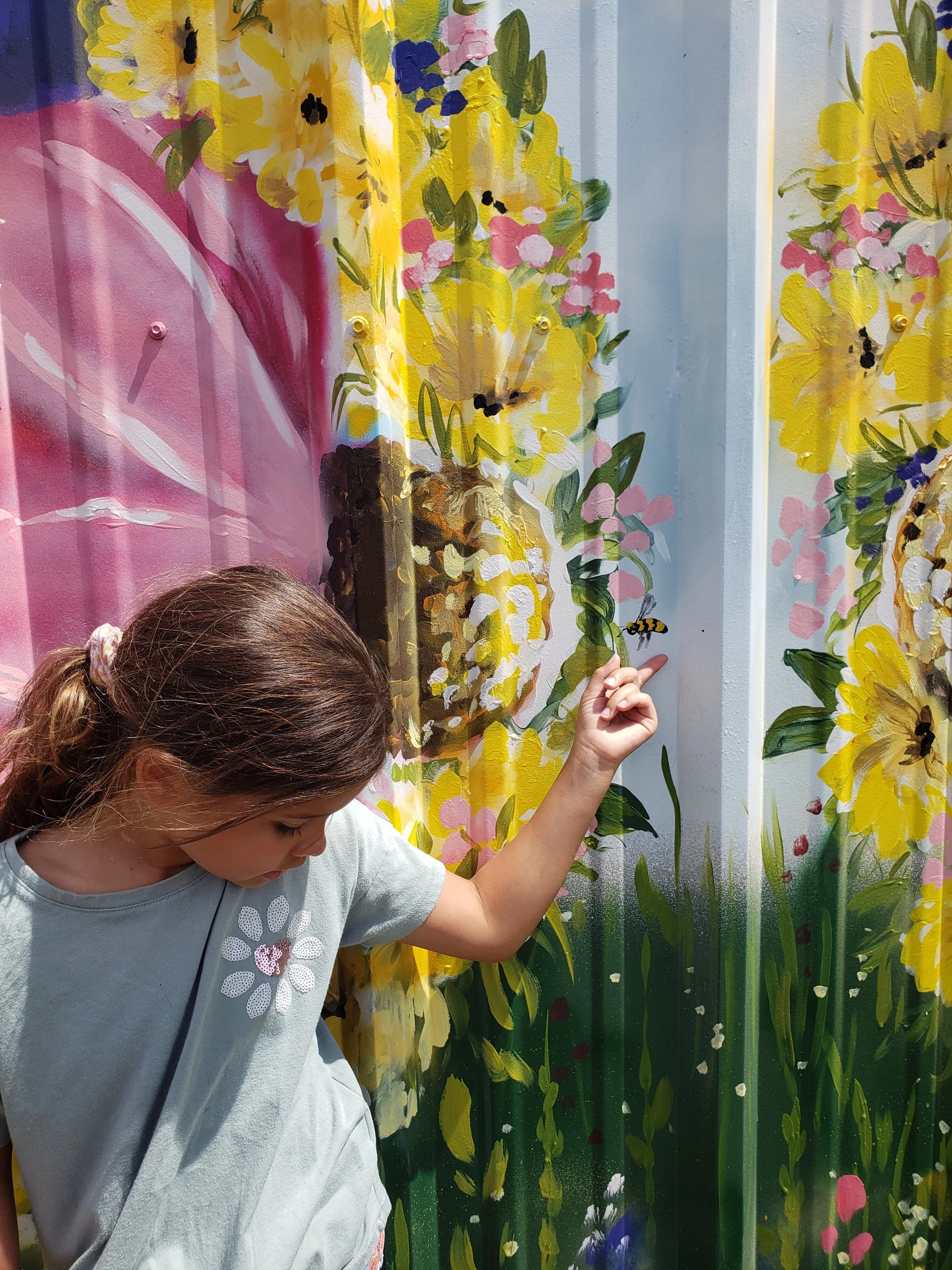 Young girl with brown hair pinned with a pink scrunchie, wearing a gray T-shirt with floral design, touching a colorful floral mural with yellow, pink, purple flowers, and a bee.