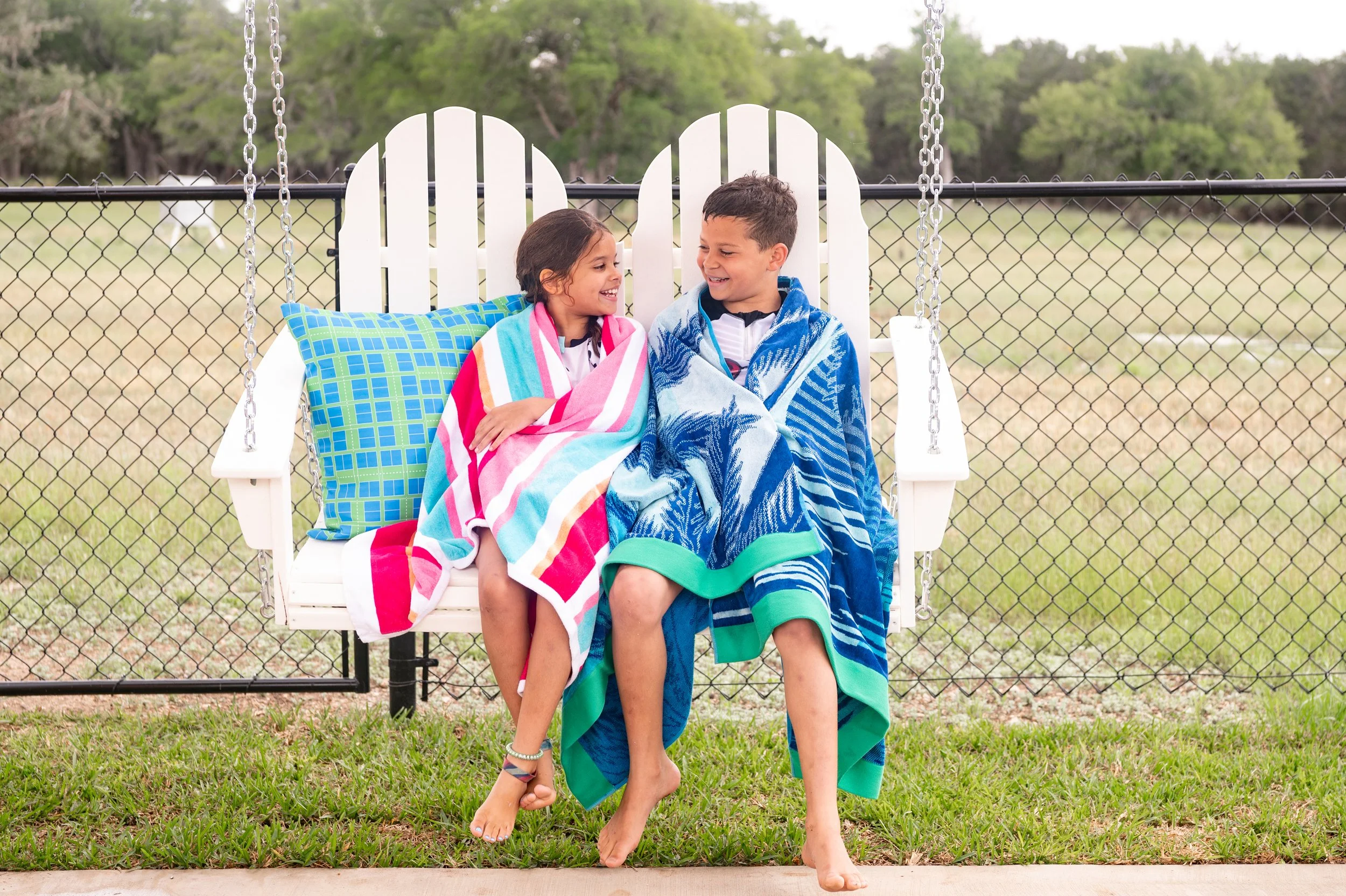 Two children wrapped in towels enjoying the swing beside the pickleball court after a swim at The Gathering in Wimberley, Texas.