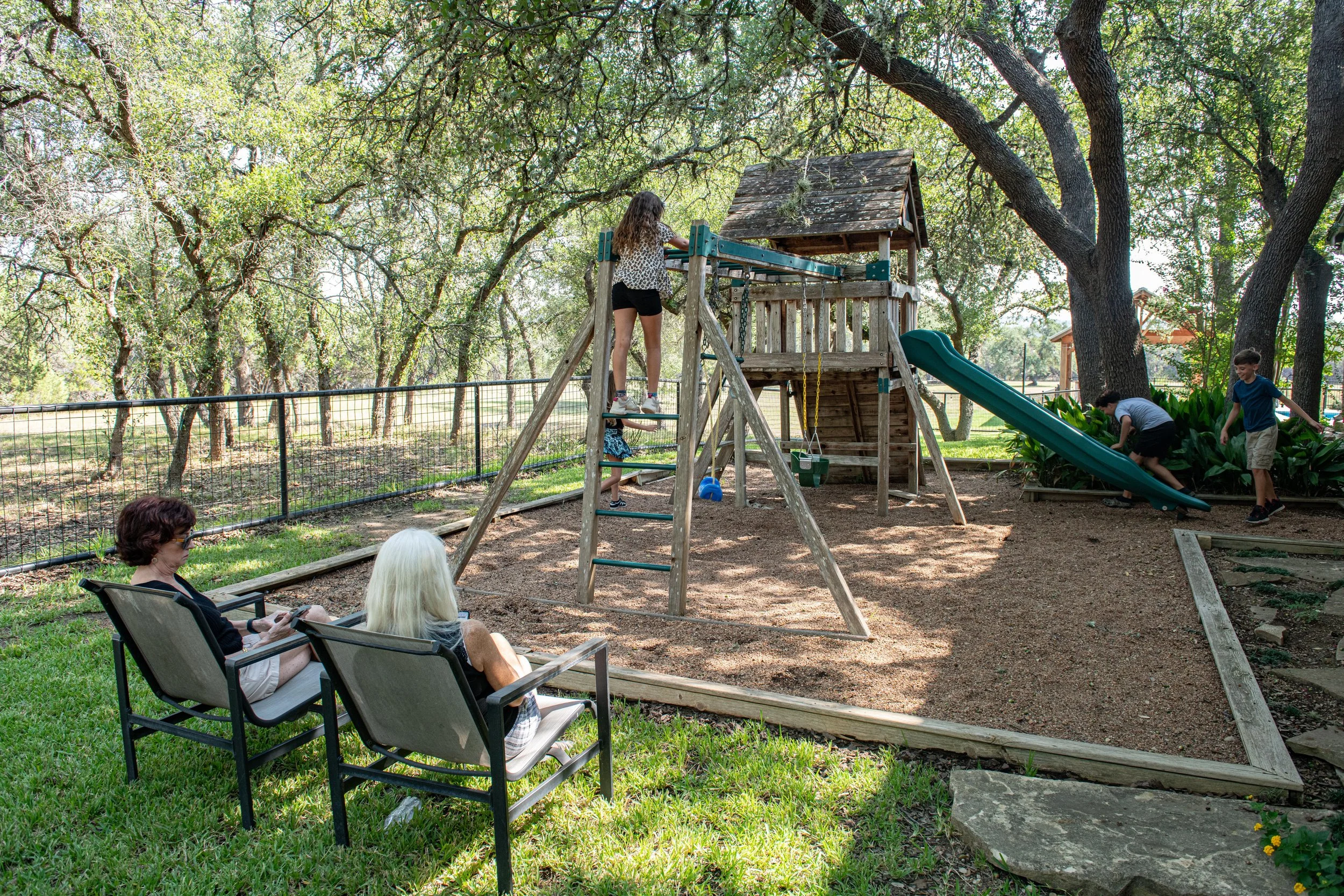 Children playing on the playground while an adult relaxes nearby under the oak trees at The Gathering.