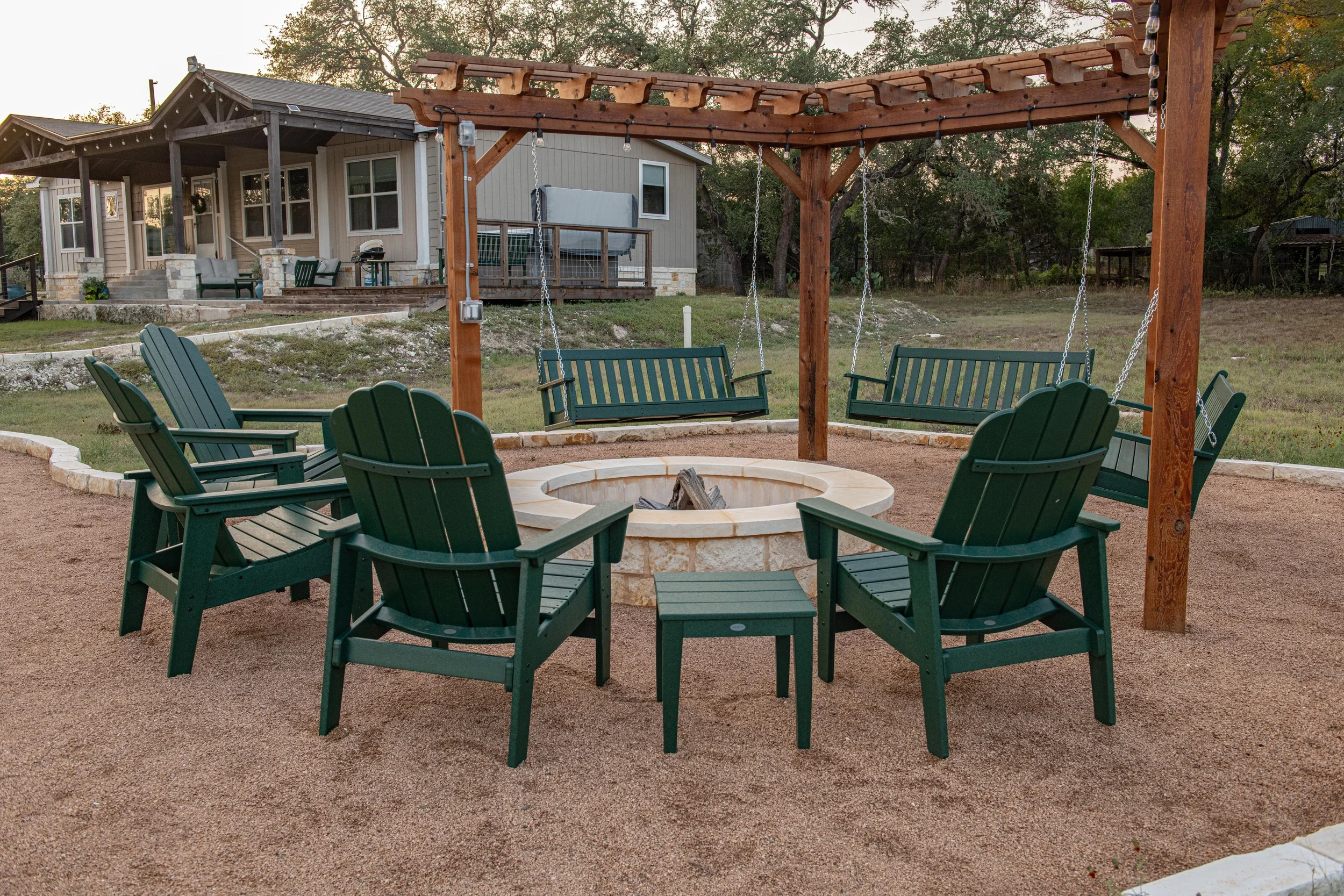 Outdoor backyard with a fire pit surrounded by six green Adirondack chairs and a wooden swing set with two swings, set on gravel with a house and trees in the background at dusk.