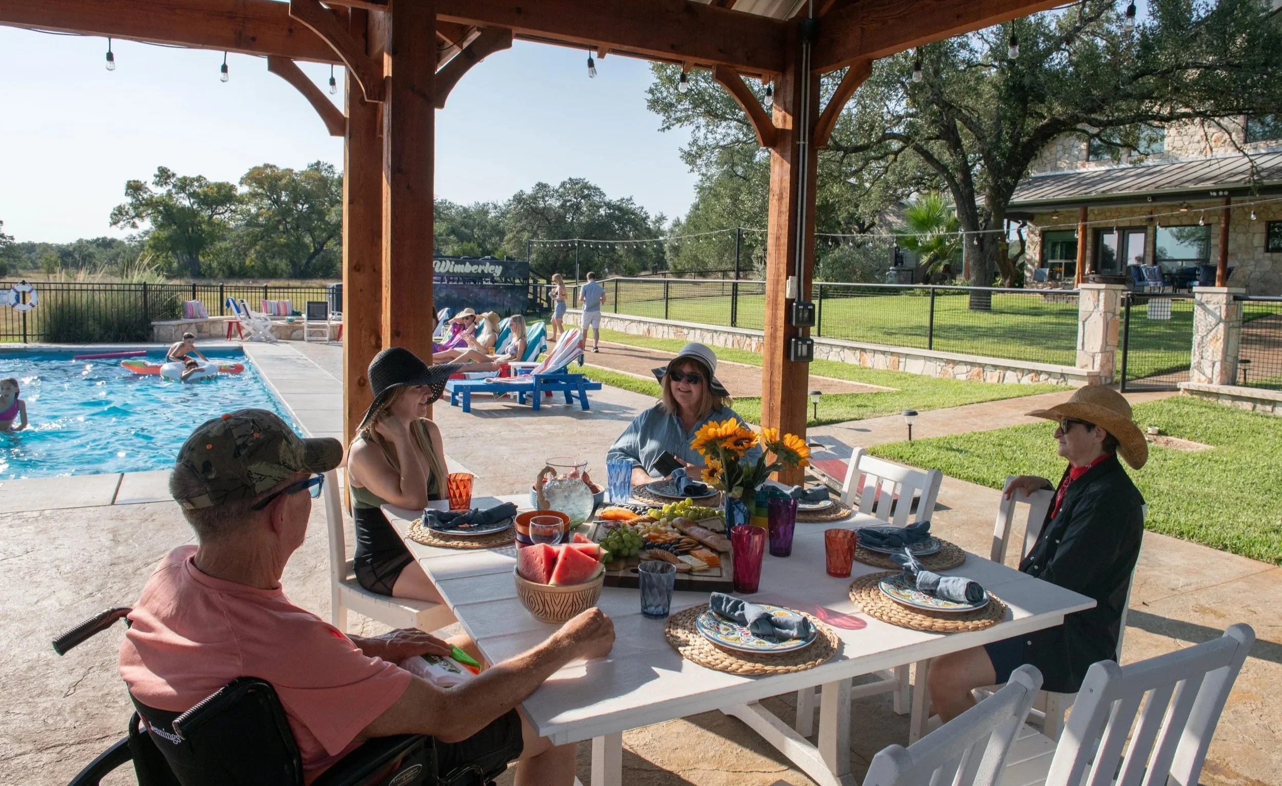 Guests of different generations sharing a meal under the pavilion while others enjoy the pool and bocce court at The Gathering in Wimberley, Texas.