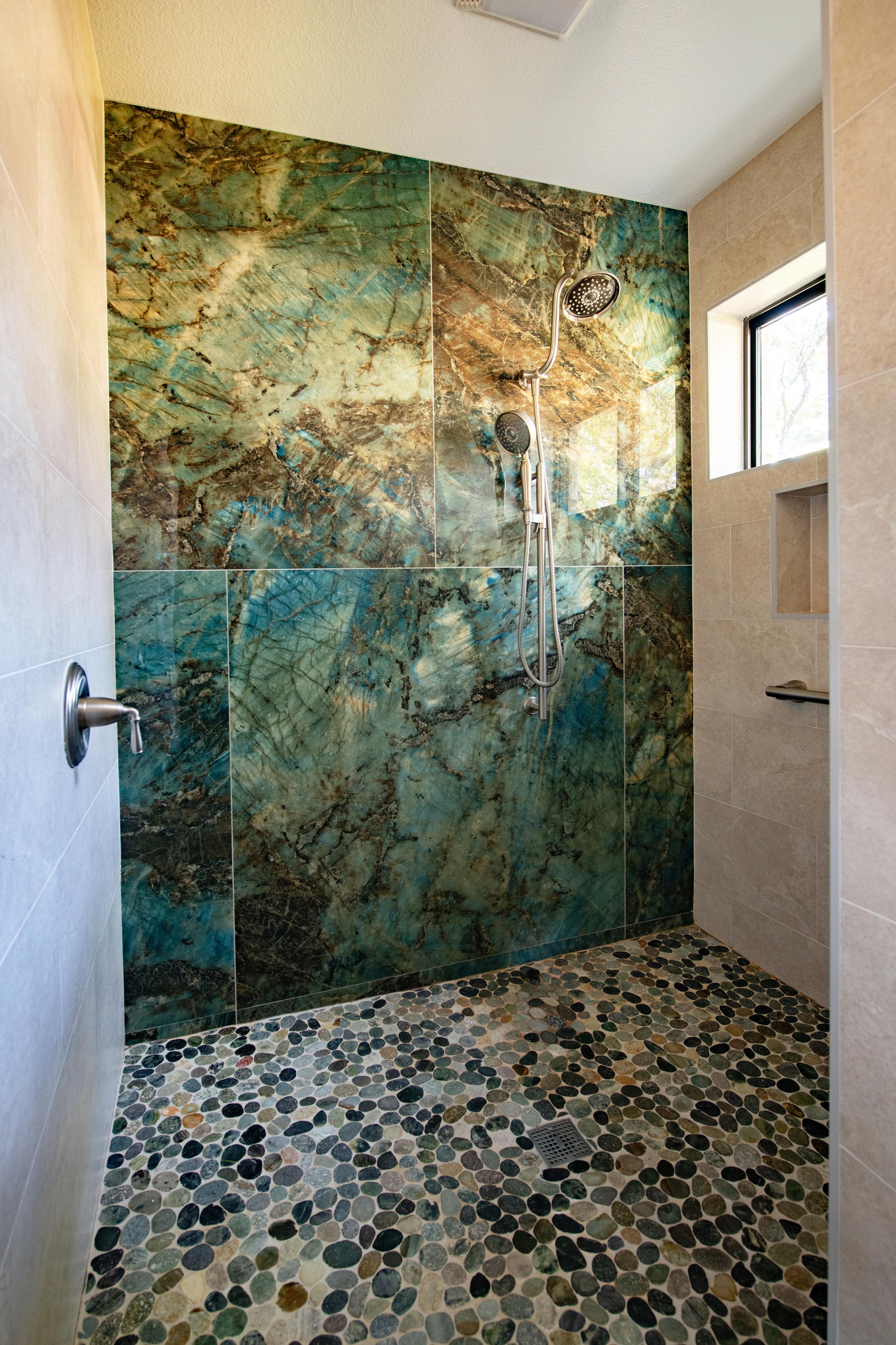 A modern walk-in shower with a pebble stone floor, large green and brown marble tiles on the back wall, a small window on the right side, and a dual shower head system.