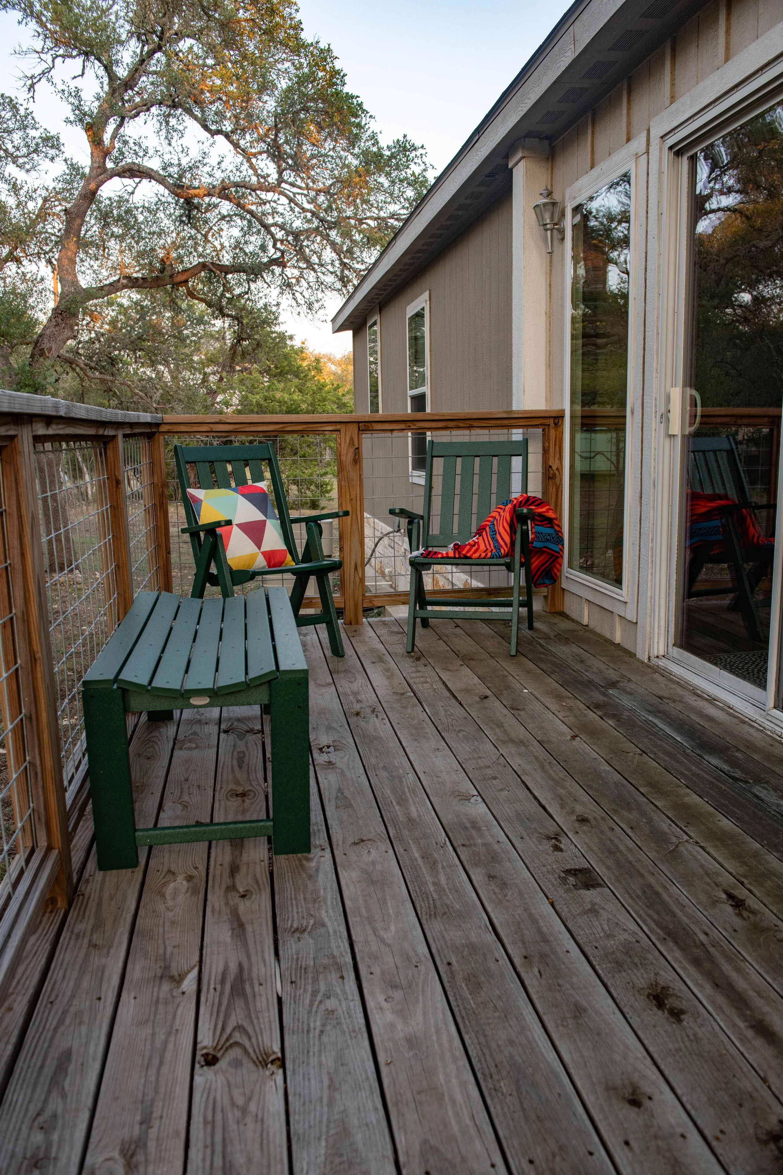 A wooden outdoor balcony or deck with two green chairs, a green bench, and a colorful pillow and blanket. There are trees in the background and a sliding glass door.