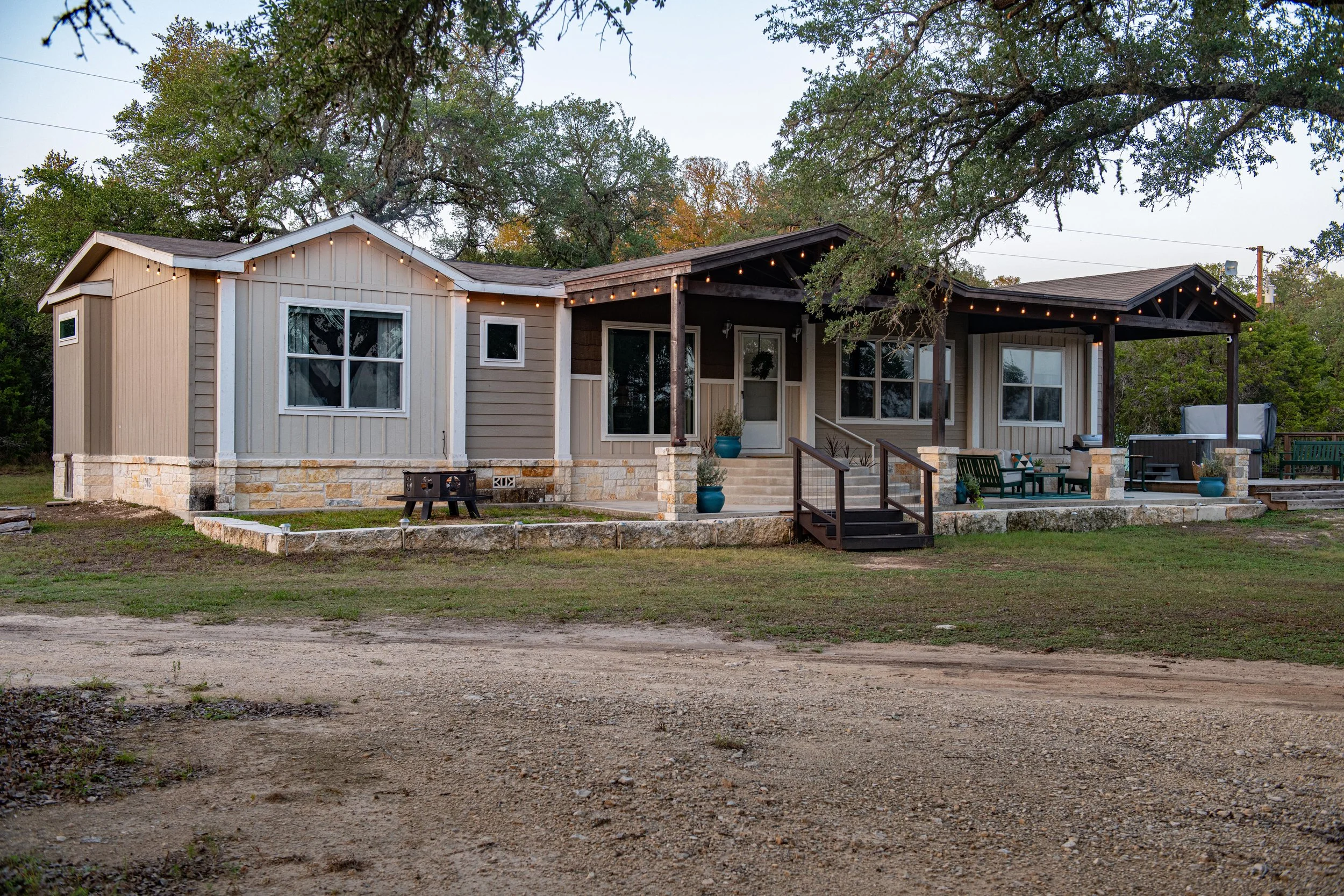 A modern single-story house with a porch, string lights, outdoor seating, potted plants, and a gravel driveway surrounded by trees.