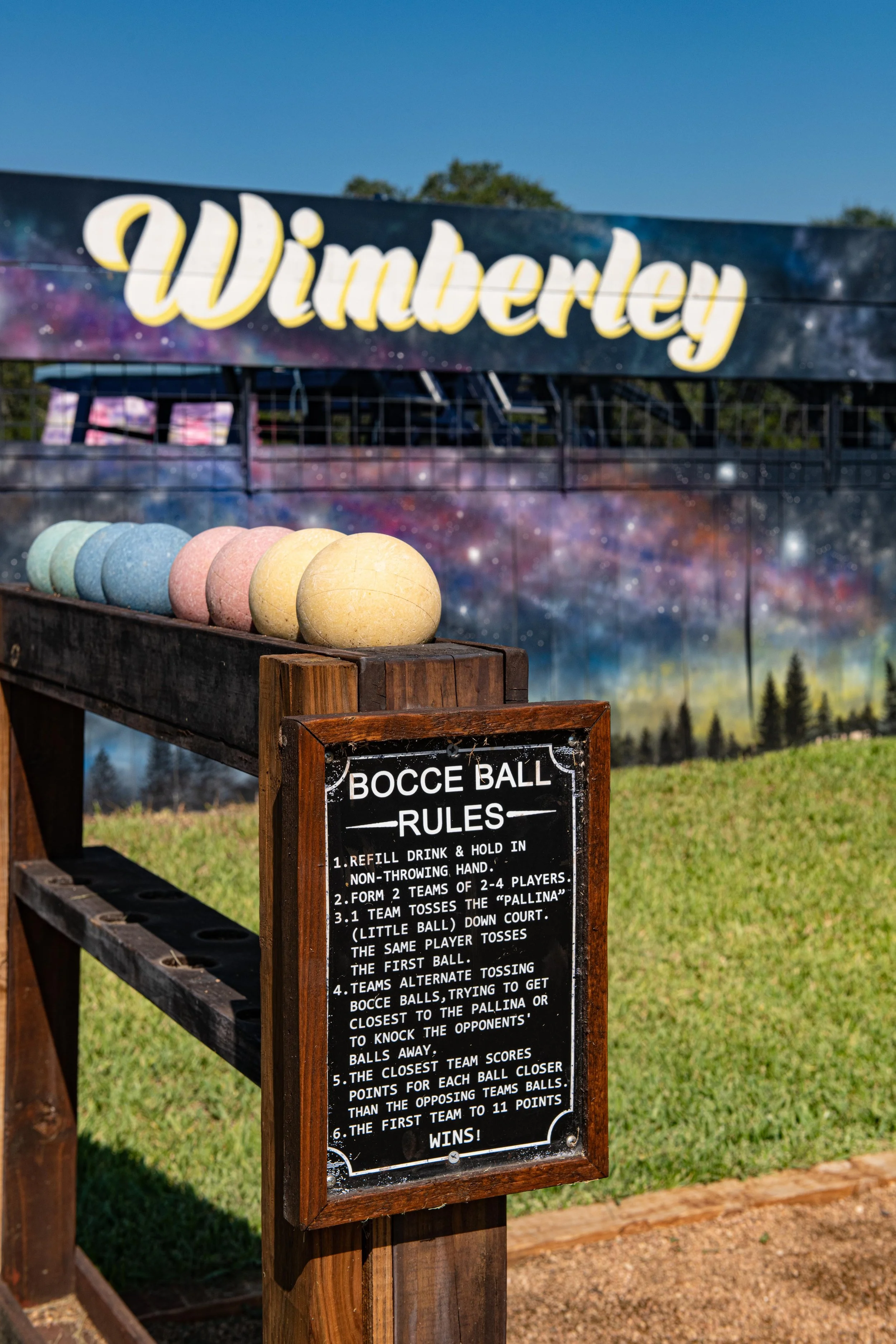 Colorful bocce balls lined up on a wooden rack with a scoreboard and a large sign reading 'Wimberley' in the background, outdoors on a sunny day.