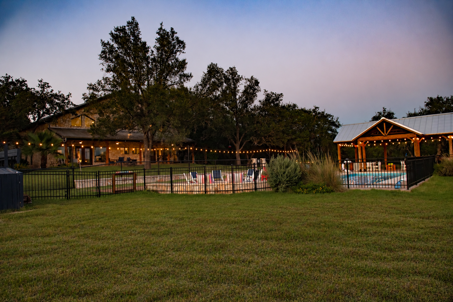 The Gathering in Wimberley, Texas at dusk, with the Main House, resort-style pool, covered pavilion, and warm string lights across the private Hill Country property.