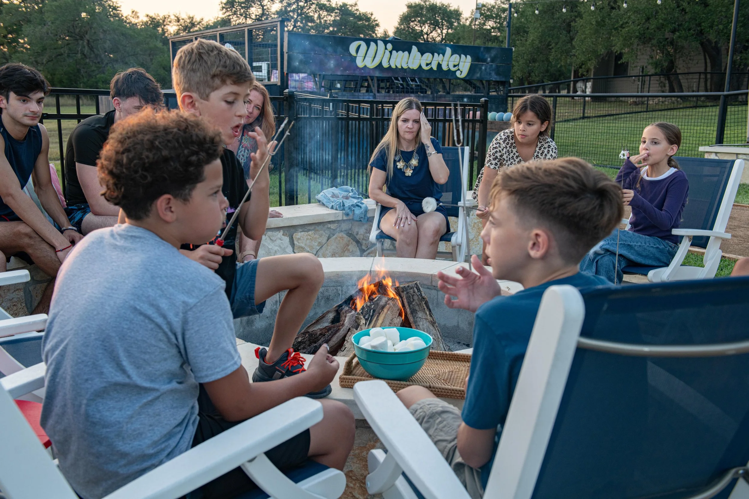 Children and adults gathered around a campfire at an outdoor backyard gathering, with a sign that reads 'Wimberley' in the background.