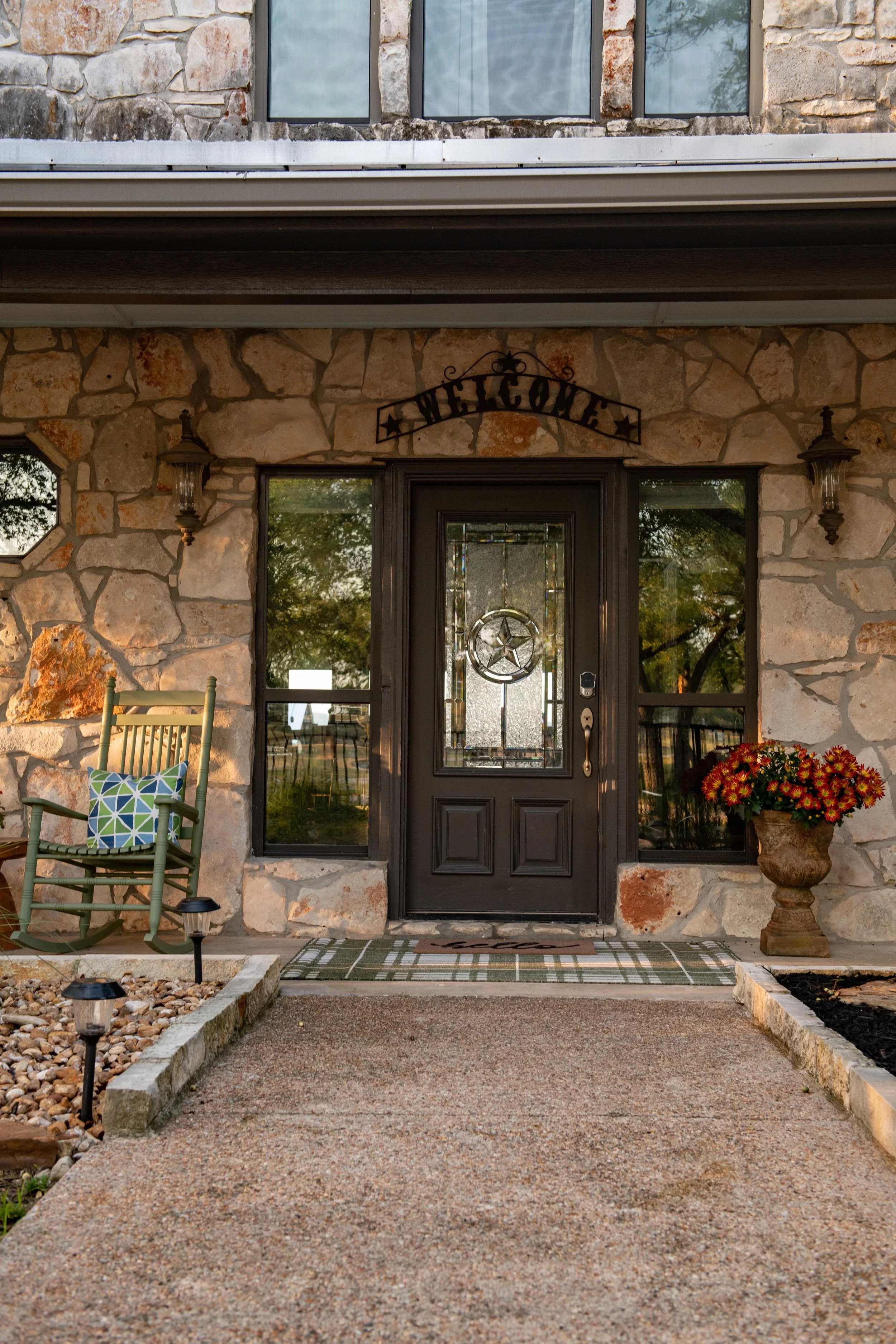 Front porch of a stone house with a black door, a welcome sign above, a rocking chair with a decorative pillow, a flower pot with red and yellow flowers, and pathway lights.