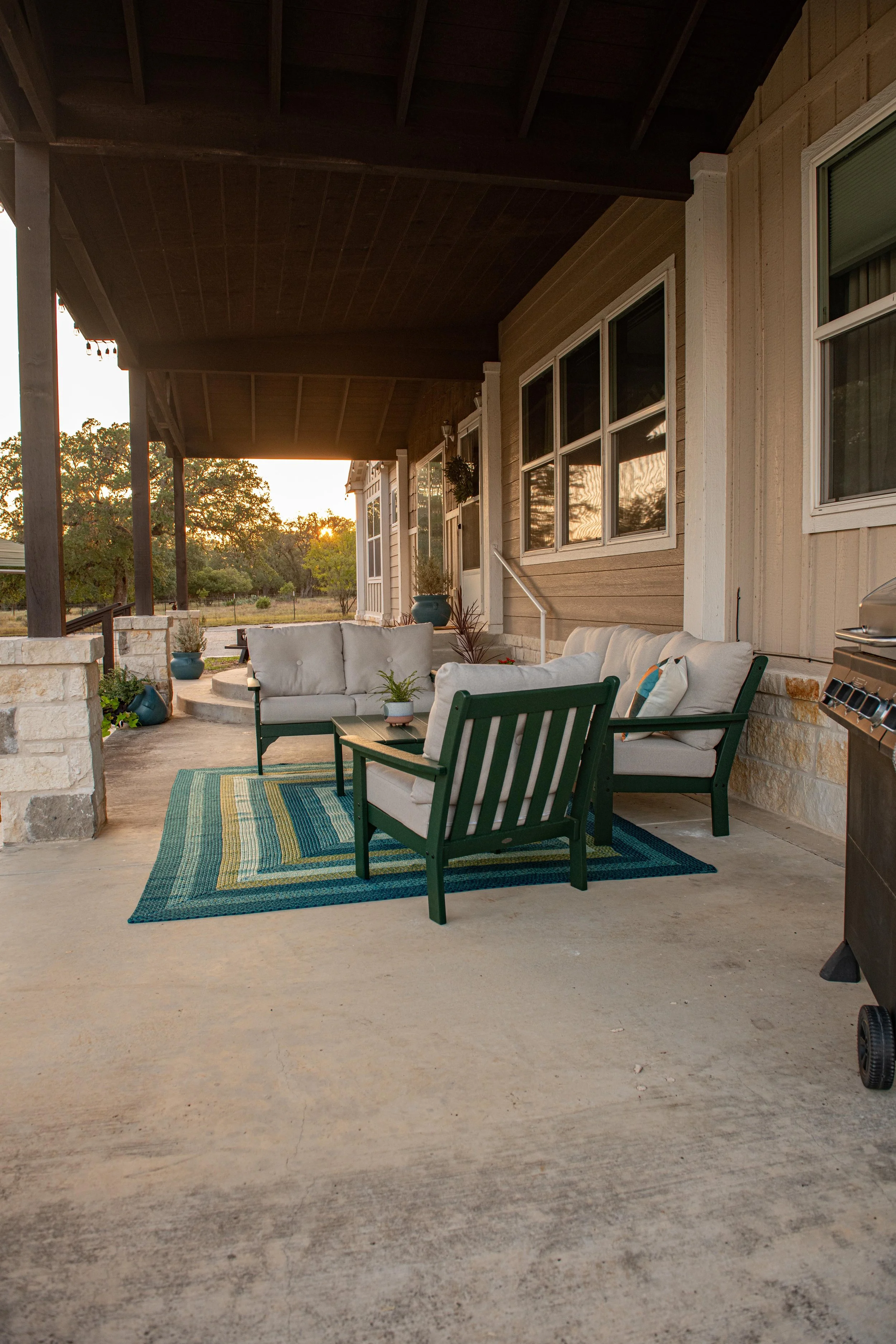 Outdoor porch with seating area including a sofa and two chairs on a patterned rug, under a wooden ceiling, with potted plants and a grill on the side, during sunset.