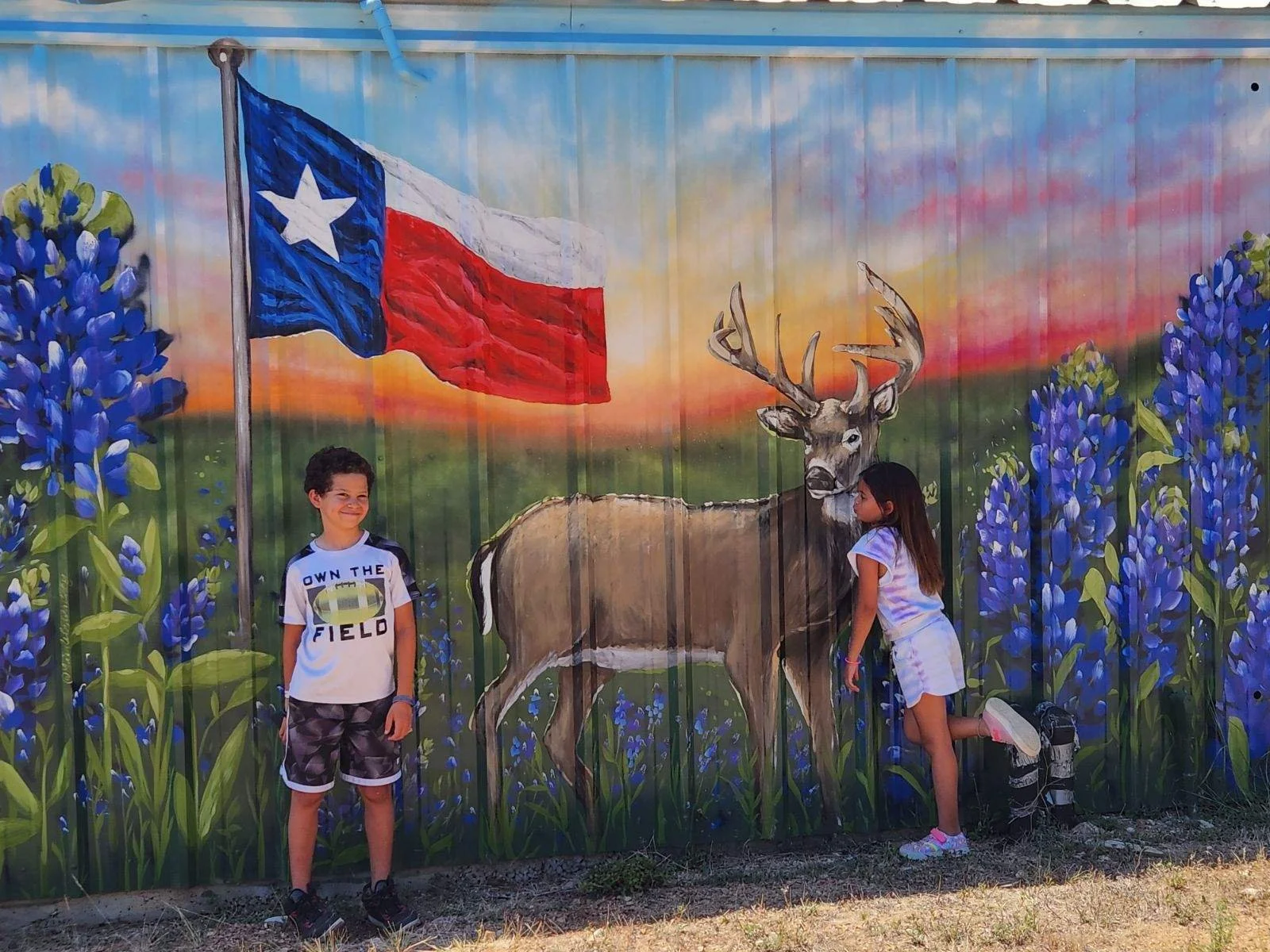 Two children standing in front of a mural with a Texas flag, a deer, and purple flowers, during sunset.
