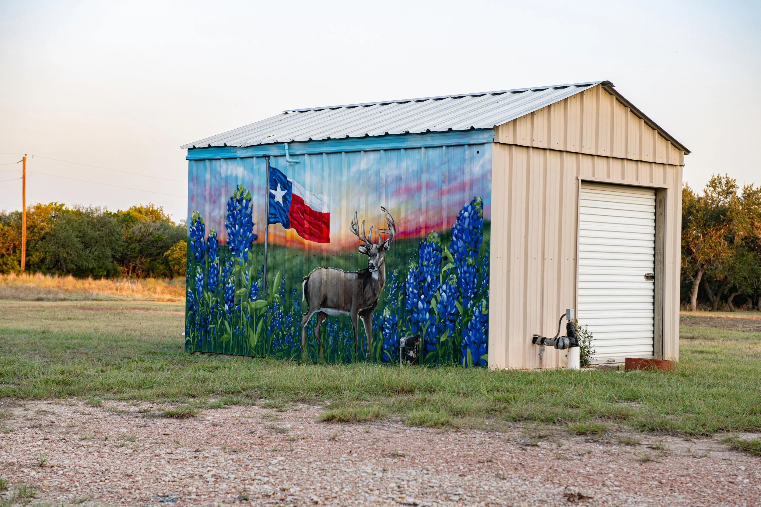 A small beige metal building with a mural on one side. The mural depicts a deer in a field of bluebonnets with the Texas flag flying in the background during sunset.