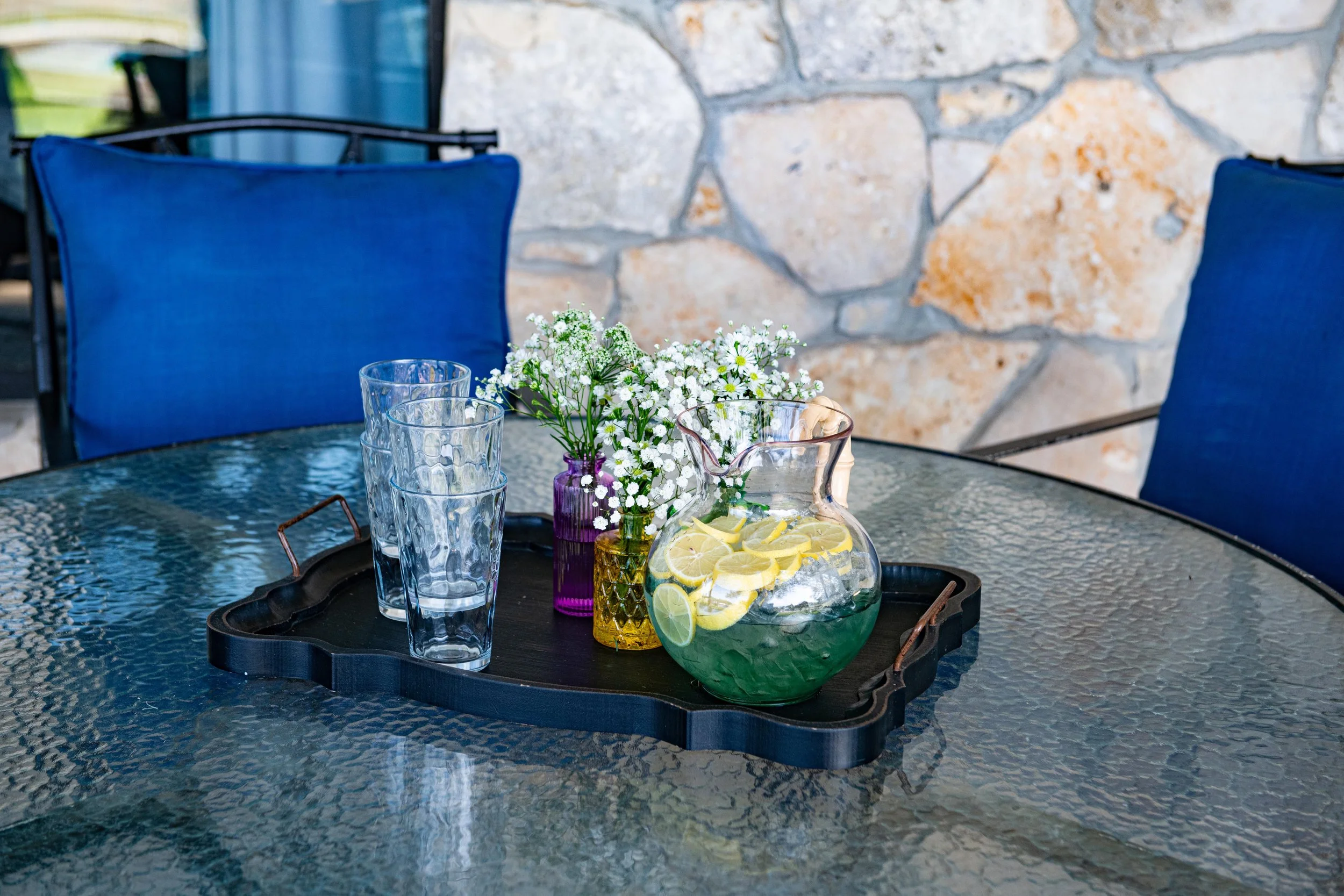 A glass outdoor table with a black tray holding a pitcher of lemon-infused green drink, three empty glasses, and two small vases with white flowers, set against a stone wall and blue patio chairs.