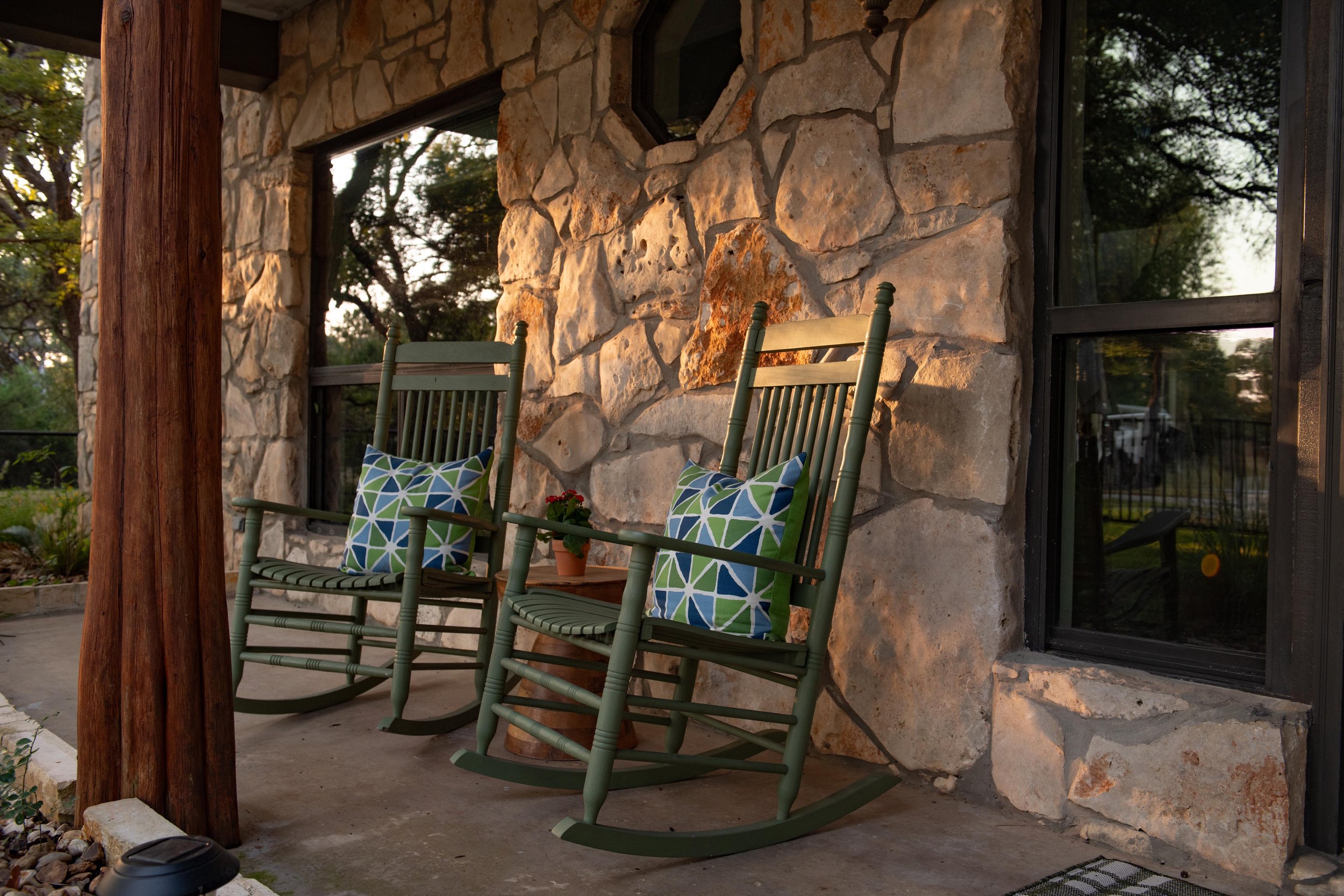 Two green wooden rocking chairs with blue and green patterned pillows are on a porch with a stone wall and large windows; a small potted plant is between the chairs, and a wooden support post is on the left.