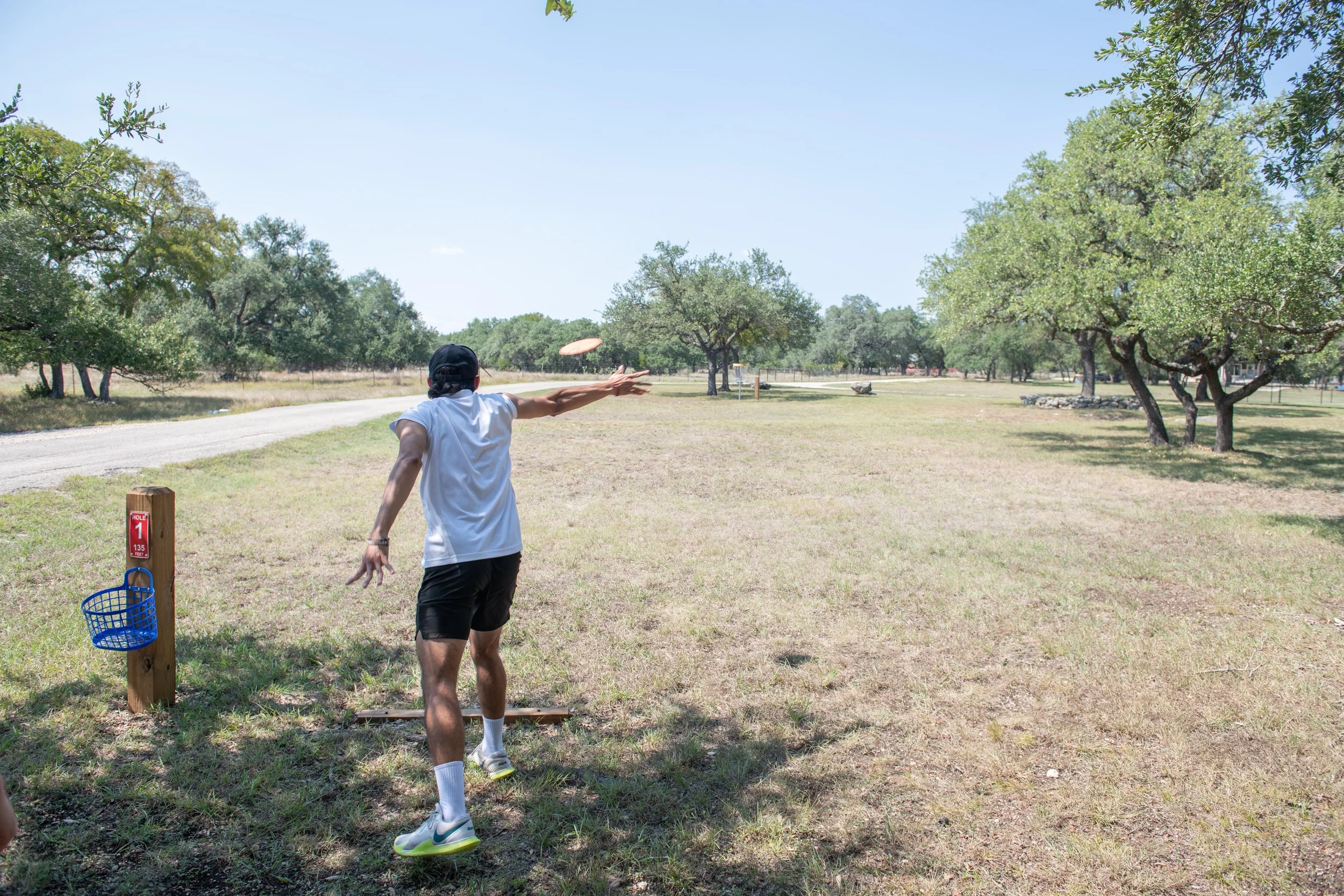 A person in a white T-shirt and black shorts playing disc golf outdoors on a sunny day, throwing a disc towards a target, with trees and a gravel path in the background.