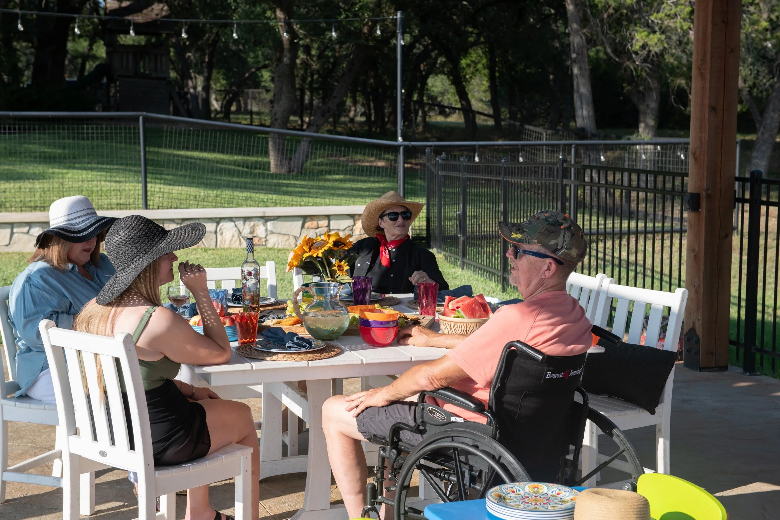 A group of five people having a meal outdoors at a patio table, with one man in a wheelchair, surrounded by watermelon, bowls, and drinks, under a shaded area with a fenced backyard.