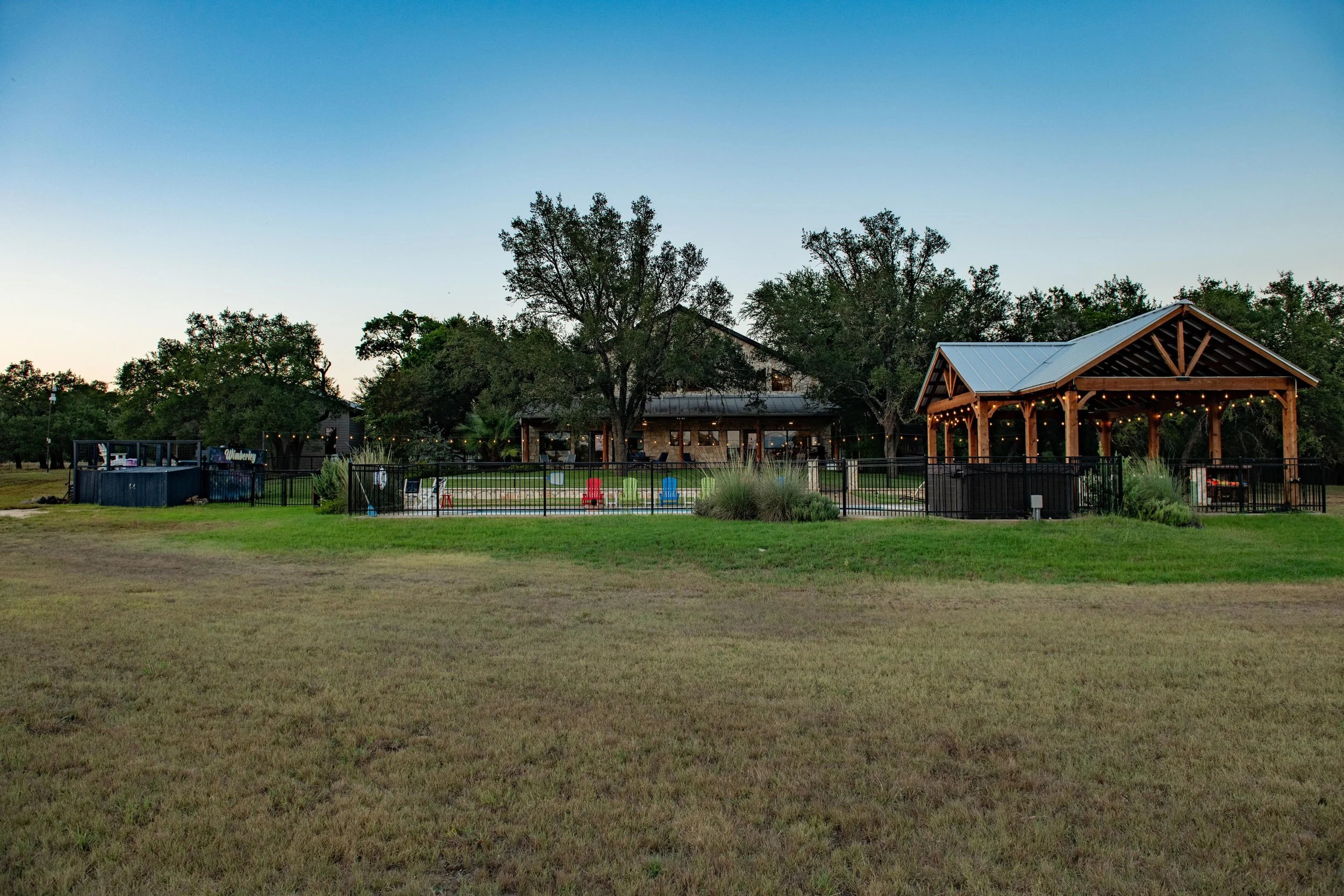 A large outdoor recreational area with a building, pavilion, and a fenced-in pool, surrounded by trees and open grassy space, during dusk.