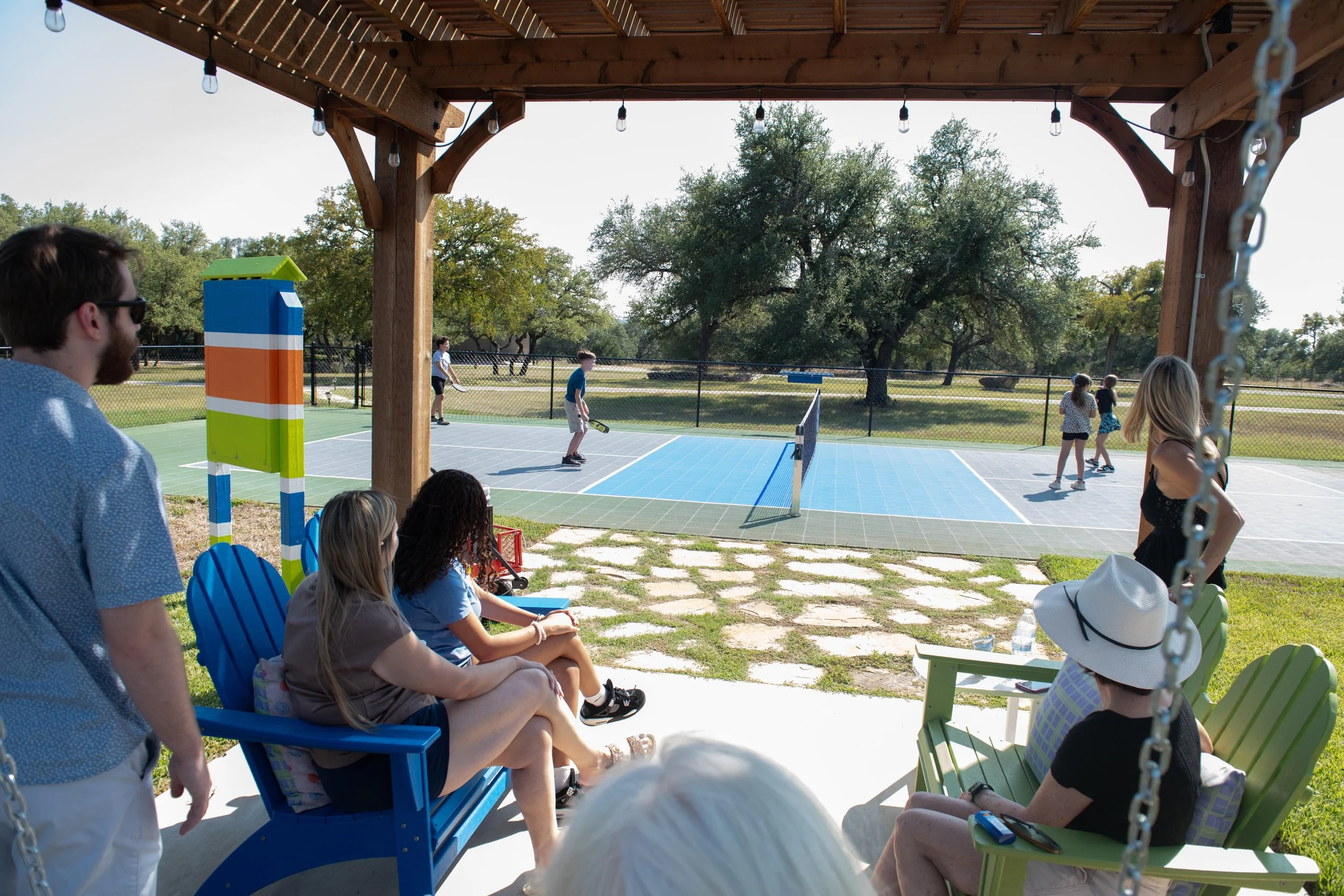 People sitting under a wooden pavilion watching children play pickleball on a court outside.