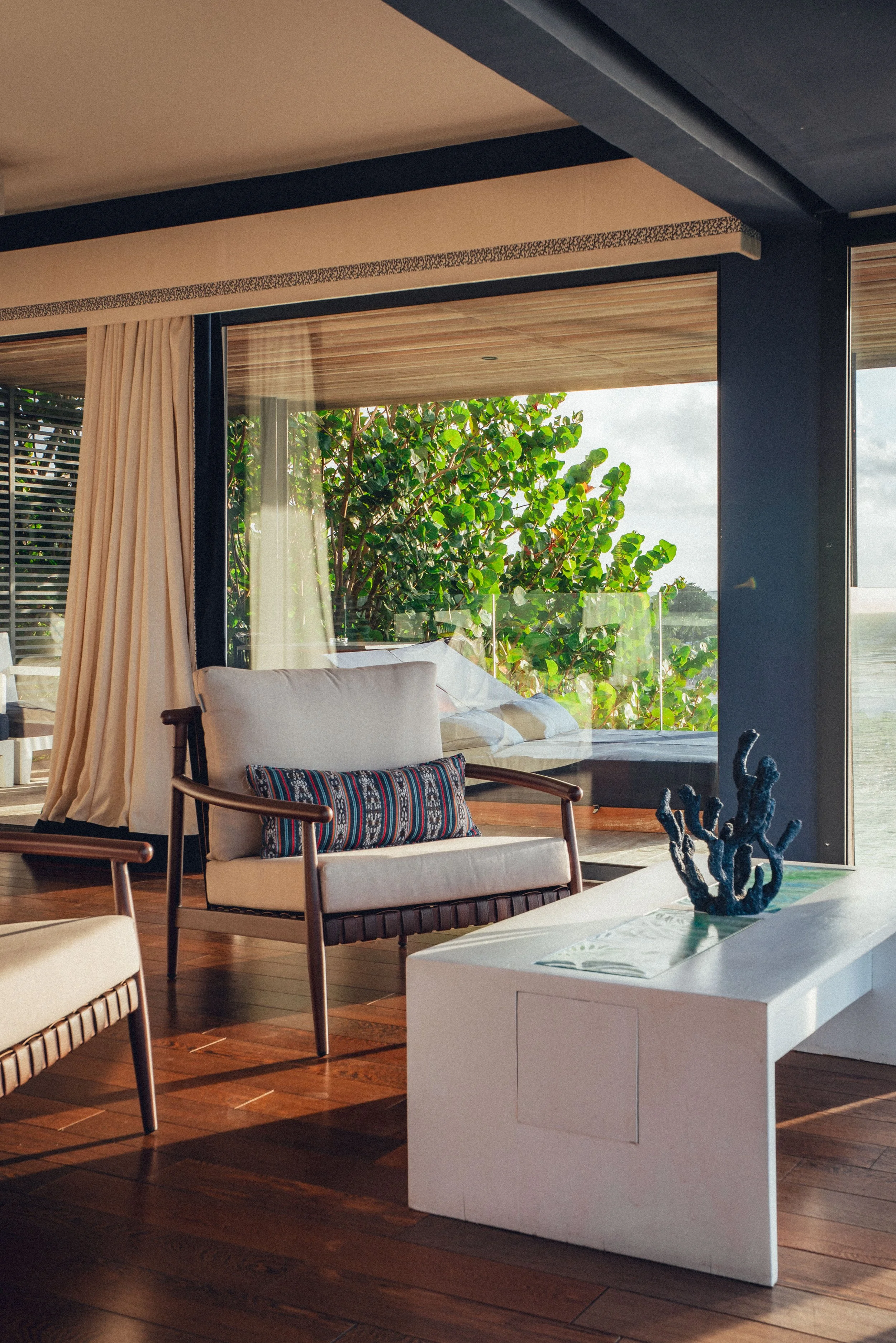 Salon intérieur avec mobilier en bois, canapé avec coussin coloré, table blanche avec décoration en forme de corail et vue sur un patio avec plantes vertes et vue extérieure.