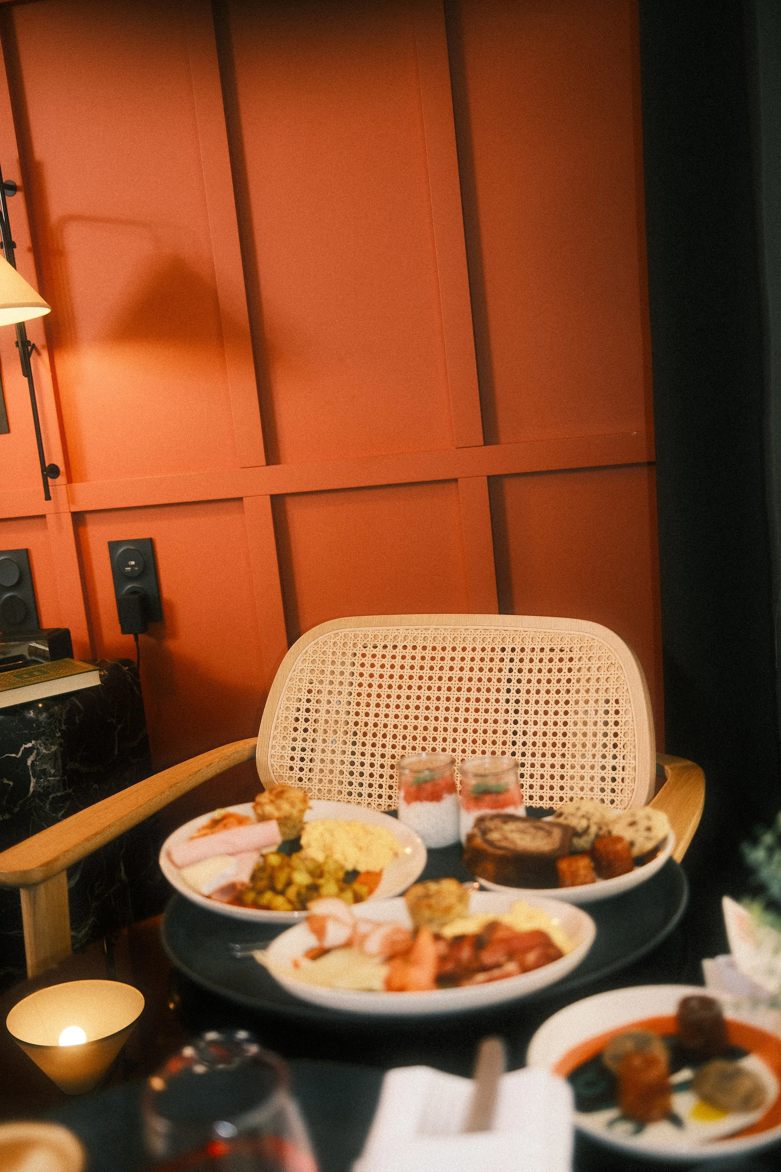 Table avec divers plats de nourriture, assiettes de sushi, desserts, et boissons, dans un cadre intérieur avec mur orange en arrière-plan.