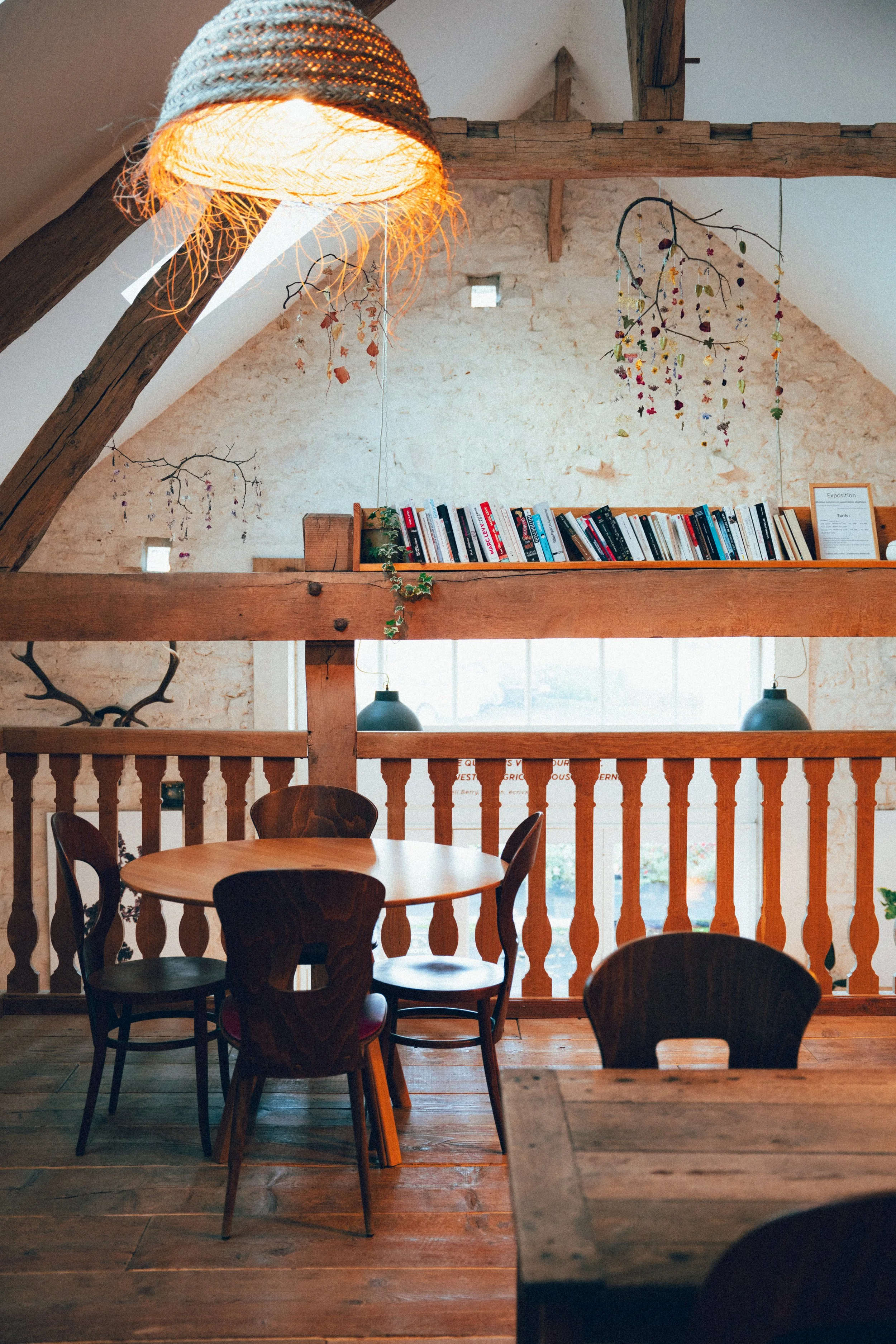 Intérieur d'un café ou d'une maison avec poutres en bois, mur en pierre, étagère avec des livres, guirlande de branches et lumières, mobilier en bois et chaises, ambiance chaleureuse et rustique.