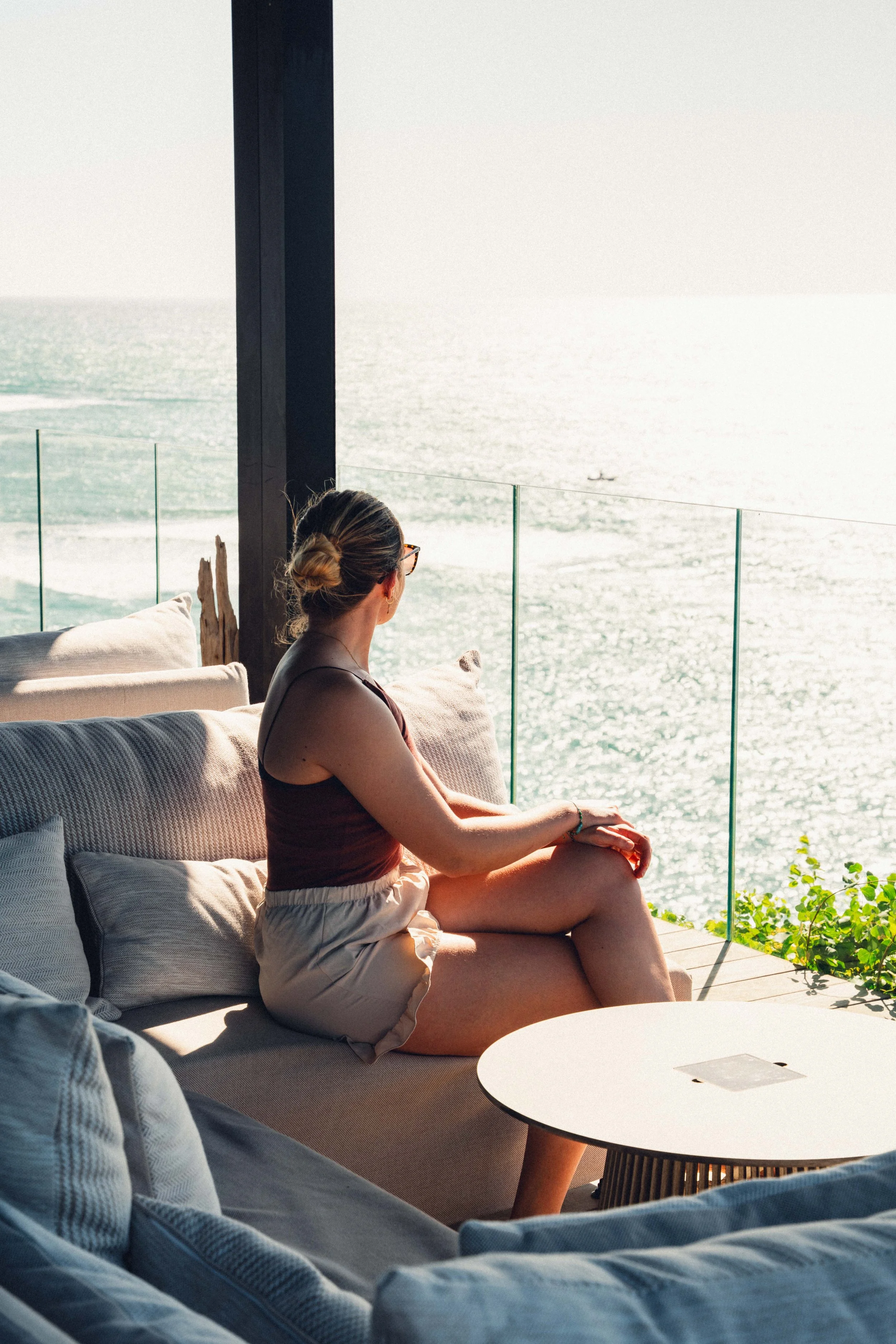Une femme assise sur un canapé près de la mer, regardant vers l'horizon, avec un verre à la main, dans un espace intérieur avec vue sur l'océan.