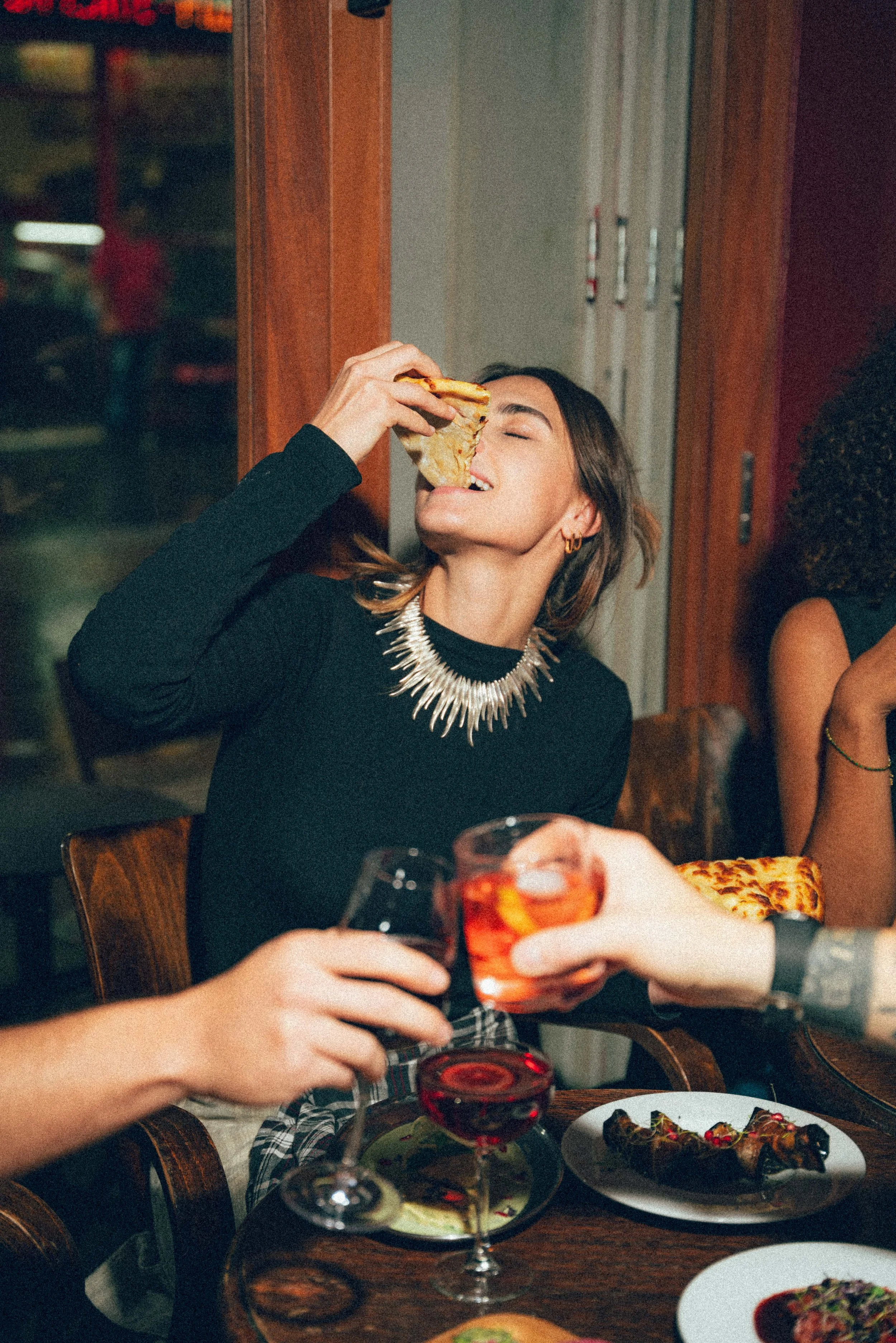 Une femme en train de manger une part de pizza dans un restaurant, d'autres personnes à la table, boissons et plats variés