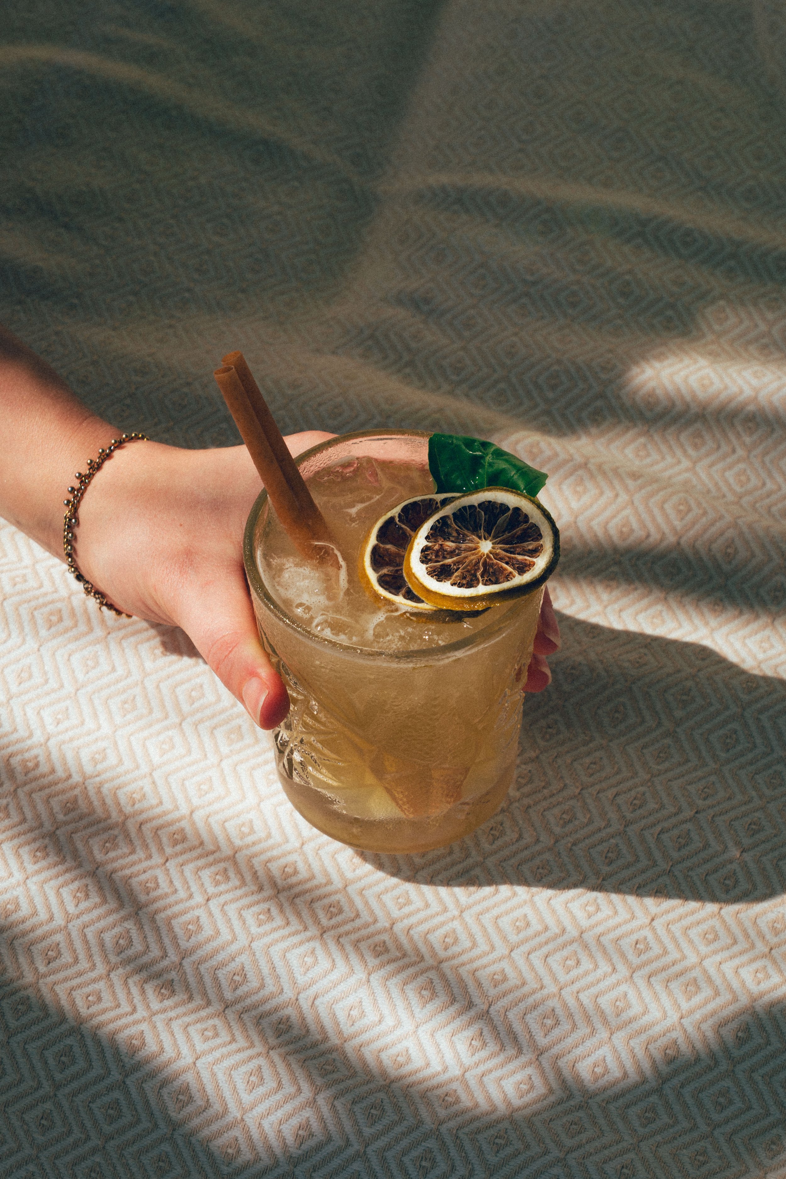 Un verre de boisson glacée garnie de rondelles de citron et de feuilles de basilic, avec une cannelle en bâton. Une main tient le verre, portant un bracelet à chaînes.