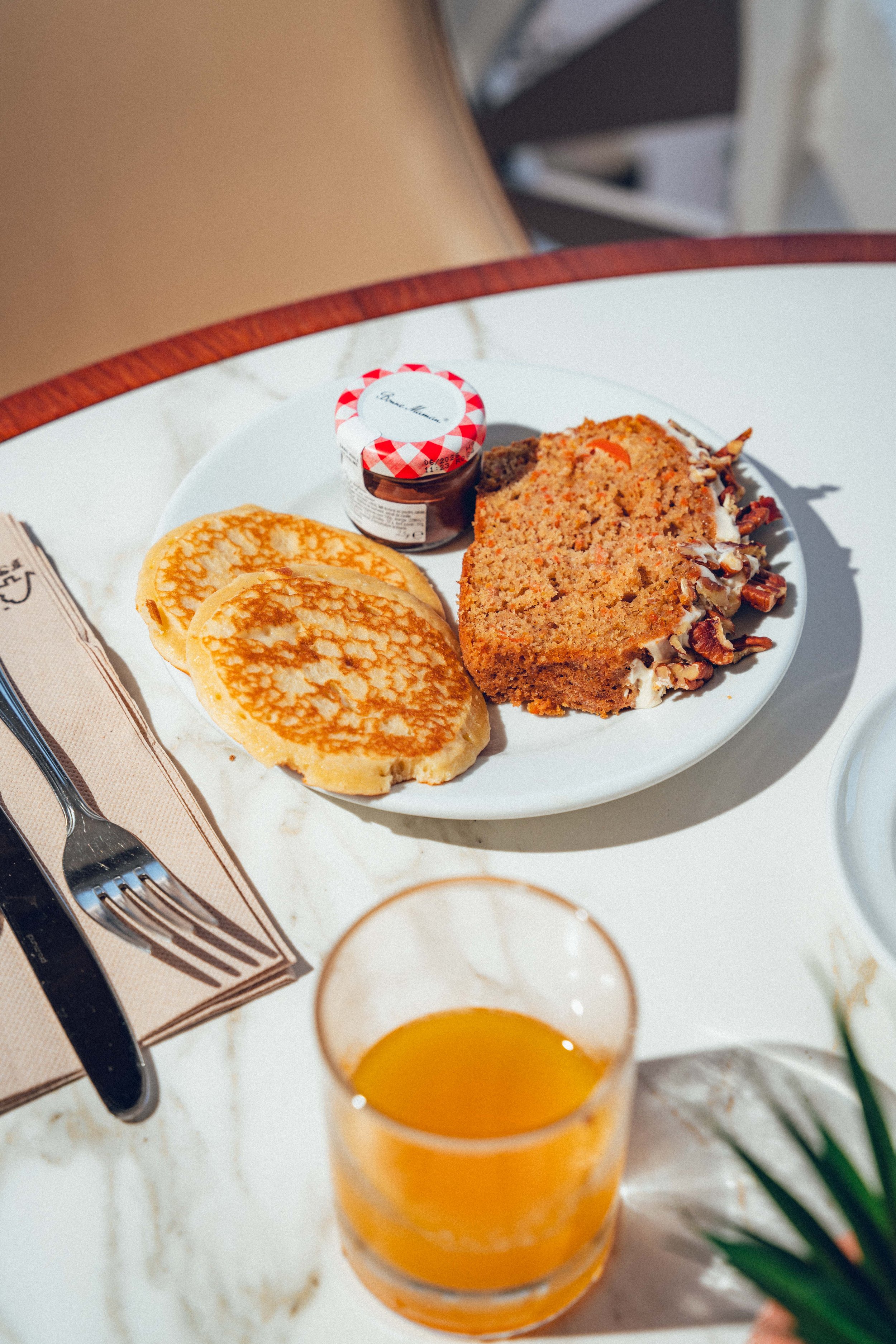Gâteau aux noix avec glaçage, pancakes, petit pot de confiture, verre de jus d'orange, nappe beige, fourchette, ciseaux, verre à vin et plante verte