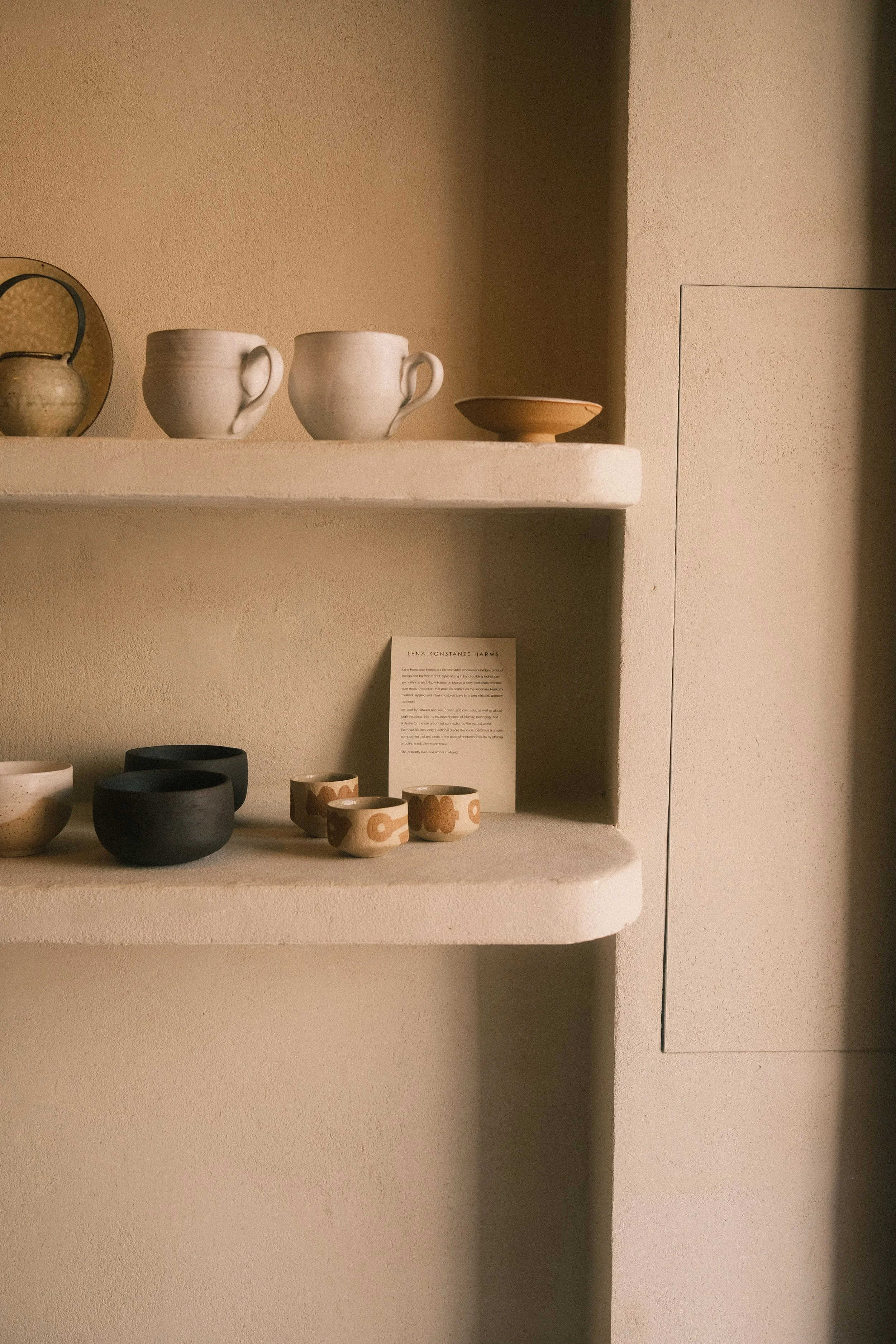 Des tasses en céramique blanches, une assiette en terre cuite et des petits bols en céramique noire et beige sont exposés sur des étagères d'une couleur beige dans une galerie d'art.