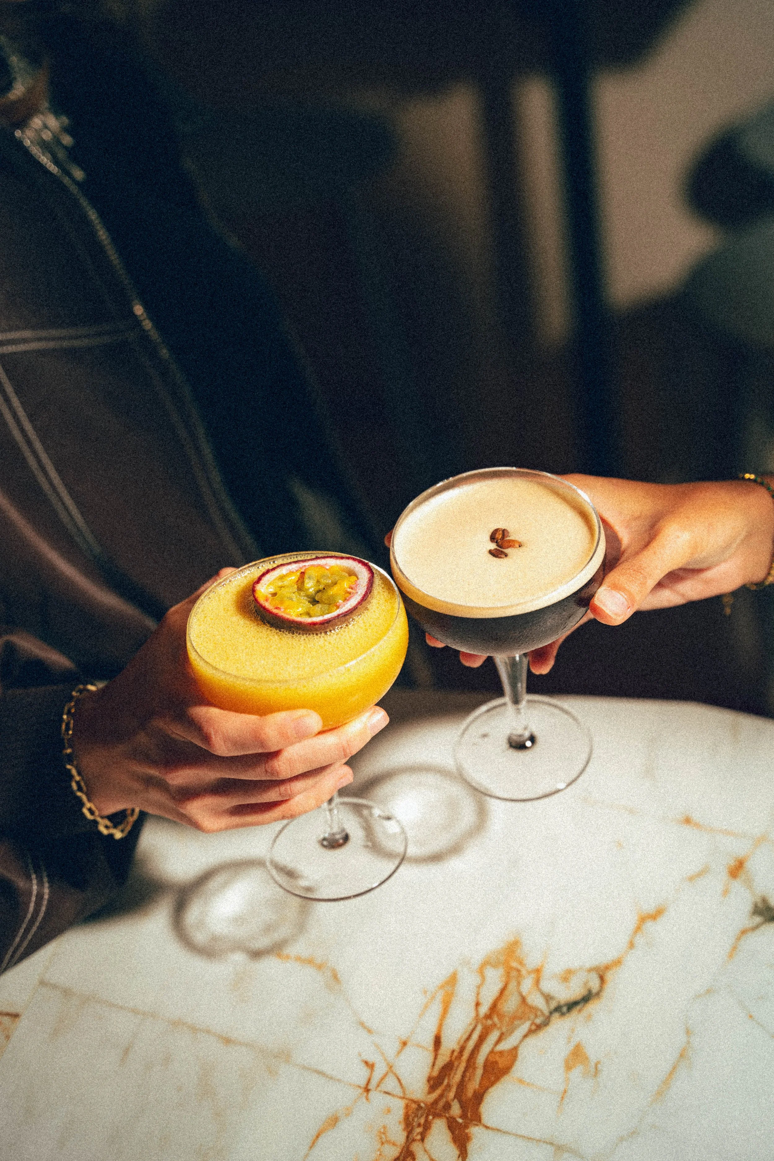 Deux personnes tiennent des cocktails dans un restaurant ou un bar, l'un avec une boisson orange décorée d'une passion ou d'un fruit, l'autre avec une boisson claire garnie de grains de café.