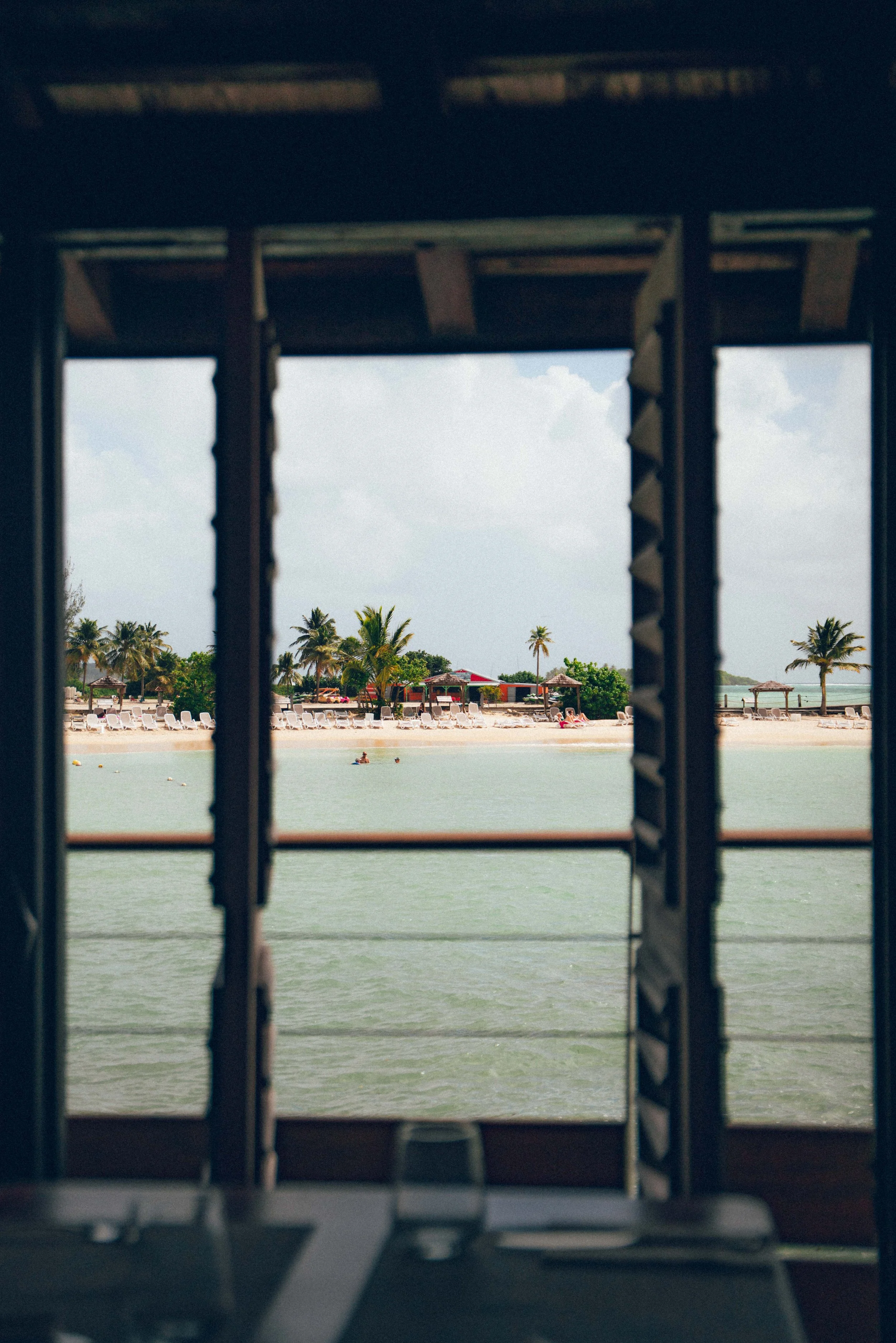Vue d'une plage avec des chaises longues, des palmiers et des cabanes, vue à travers une fenêtre en bois.