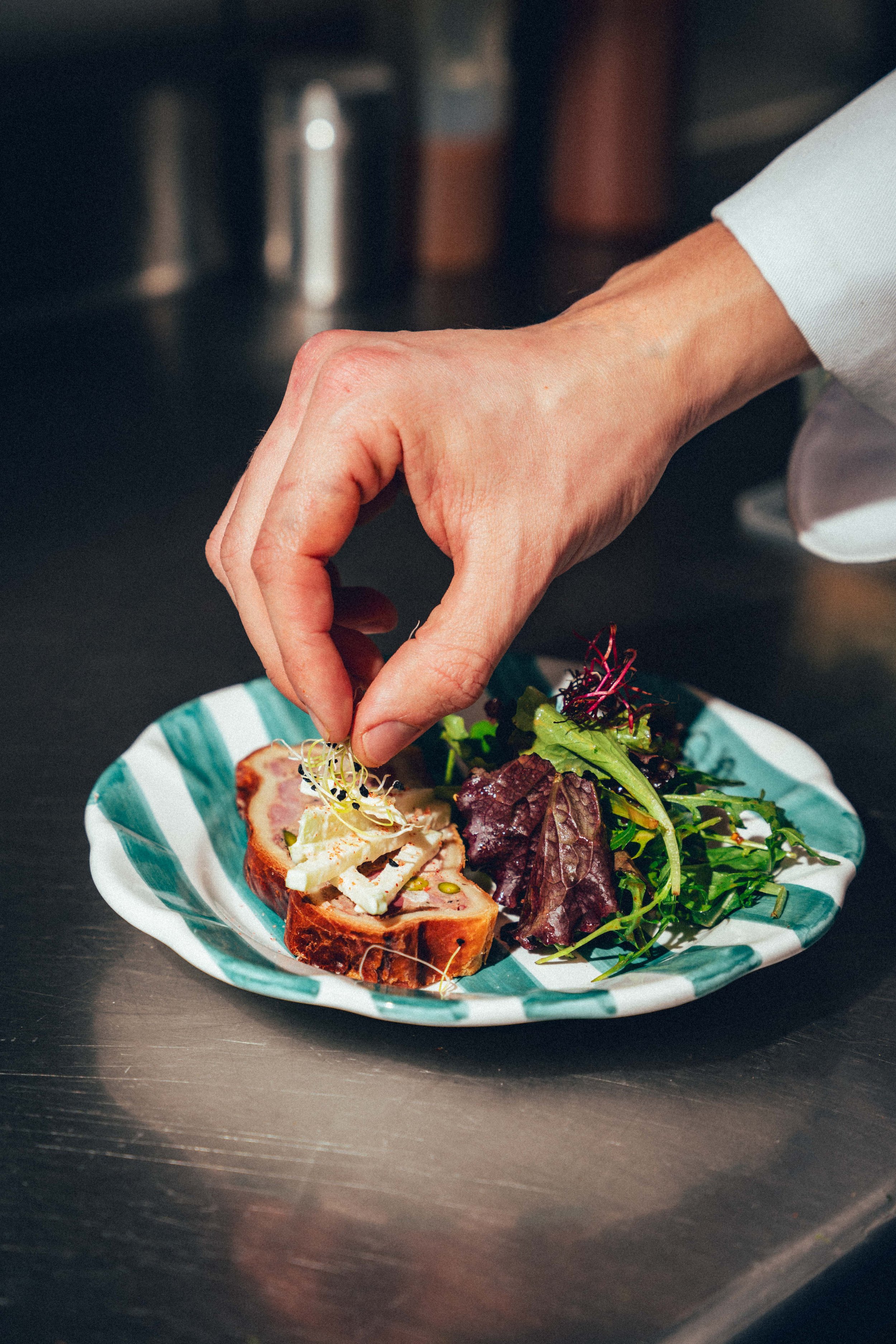 Une main déposant des micro-pousses sur une assiette contenant une tranche de pâté en croûte et une salade verte, sur une surface en métal.