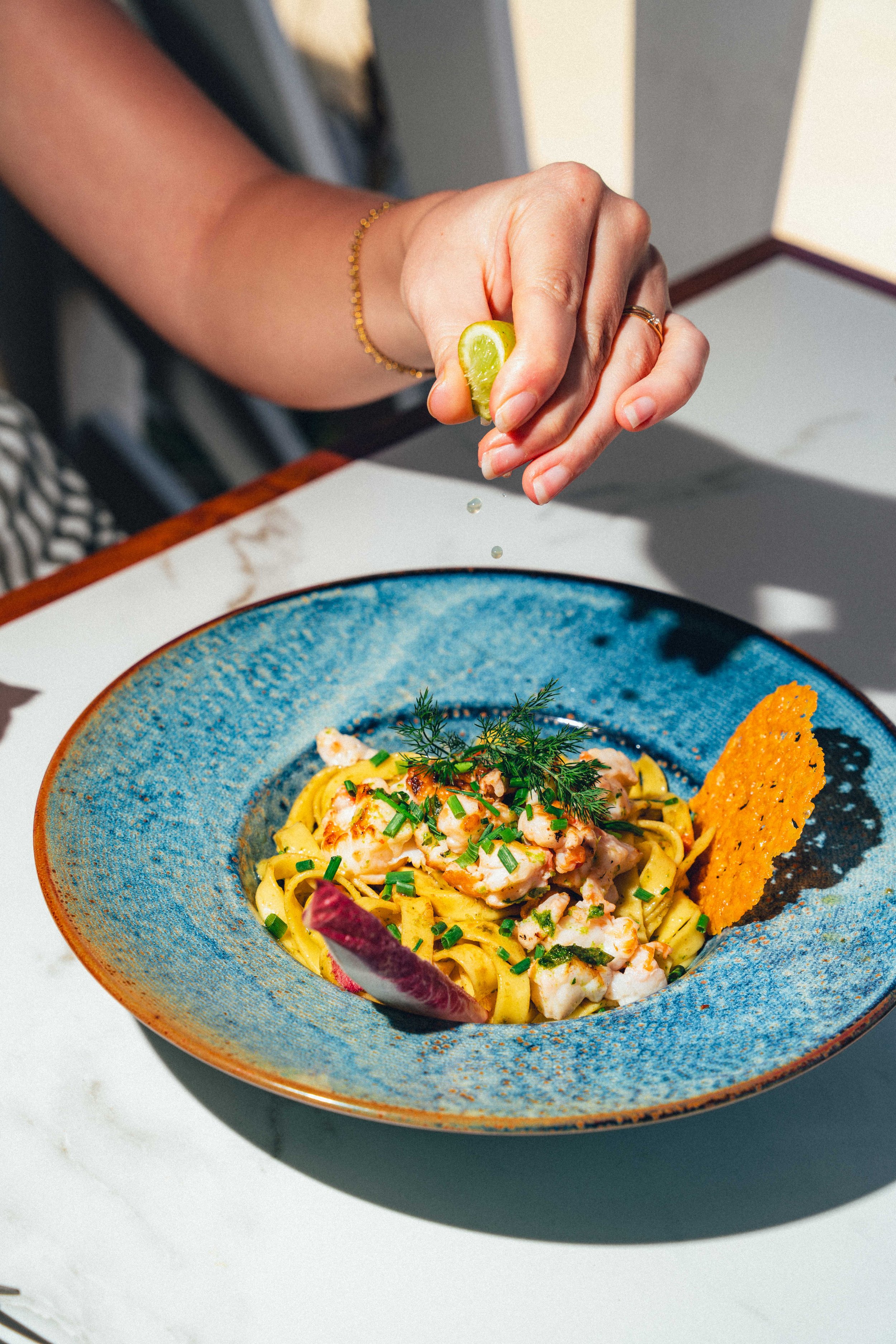 Une main tenant un morceau de citron, saupoudrant du jus sur un plat de pâtes avec des fruits de mer, servi dans une assiette bleue.