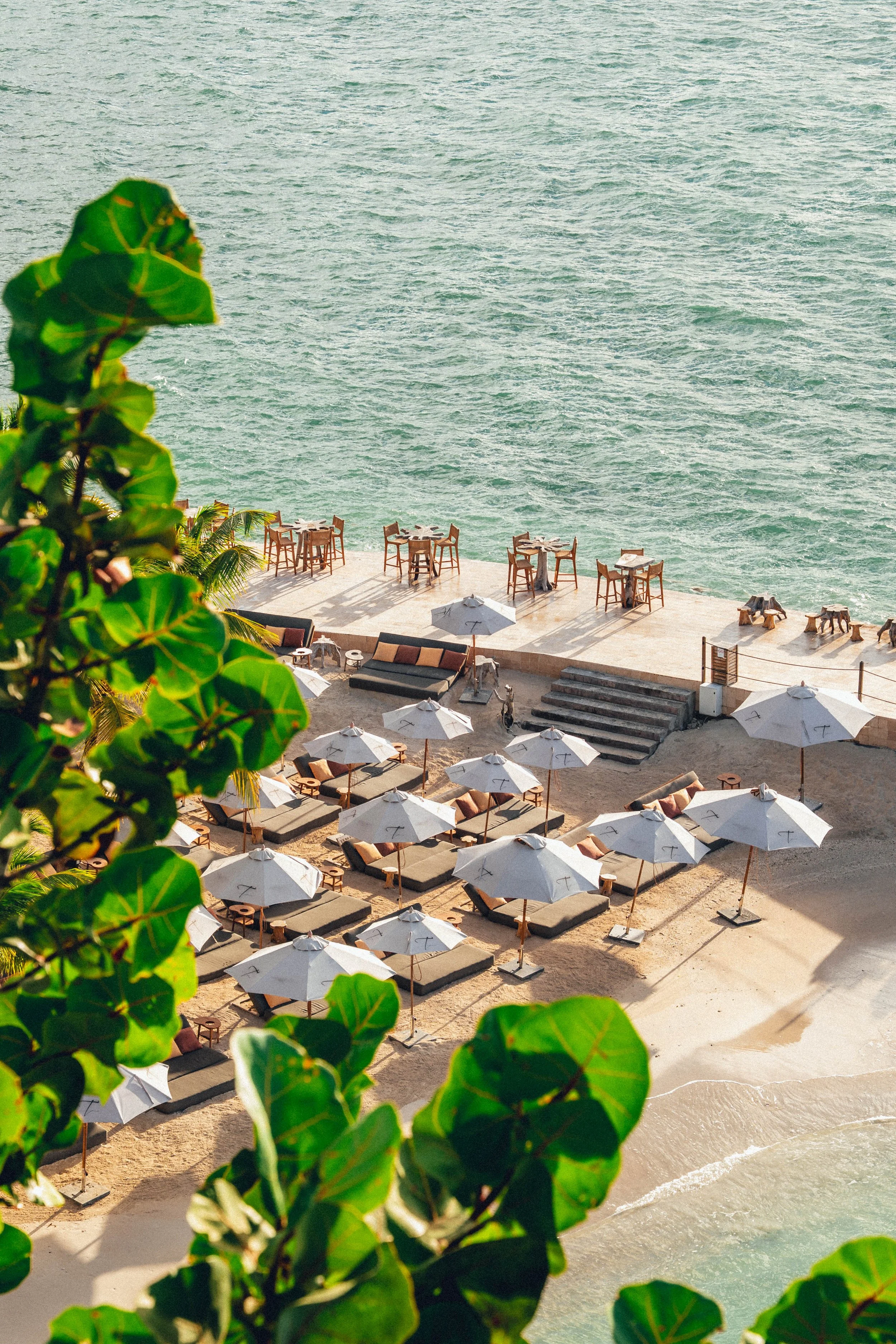 Une plage avec des parasols blancs, des lits de repos et des tables avec des chaises, vue sur la mer avec des vagues, prises d'en haut, entourée de verdure luxuriante.