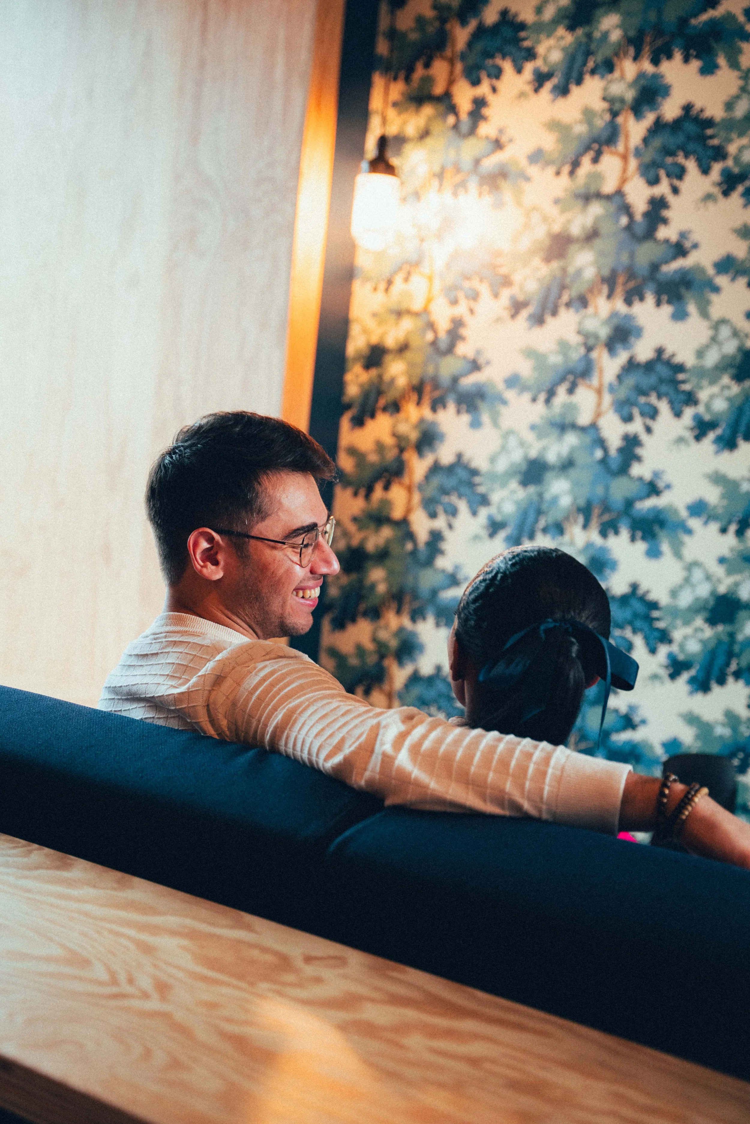 Deux personnes assises sur un canapé en train de discuter, avec un mur décoré de peinture ou de papier peint à motif de feuilles à l'arrière-plan, dans une ambiance chaleureuse et conviviale.