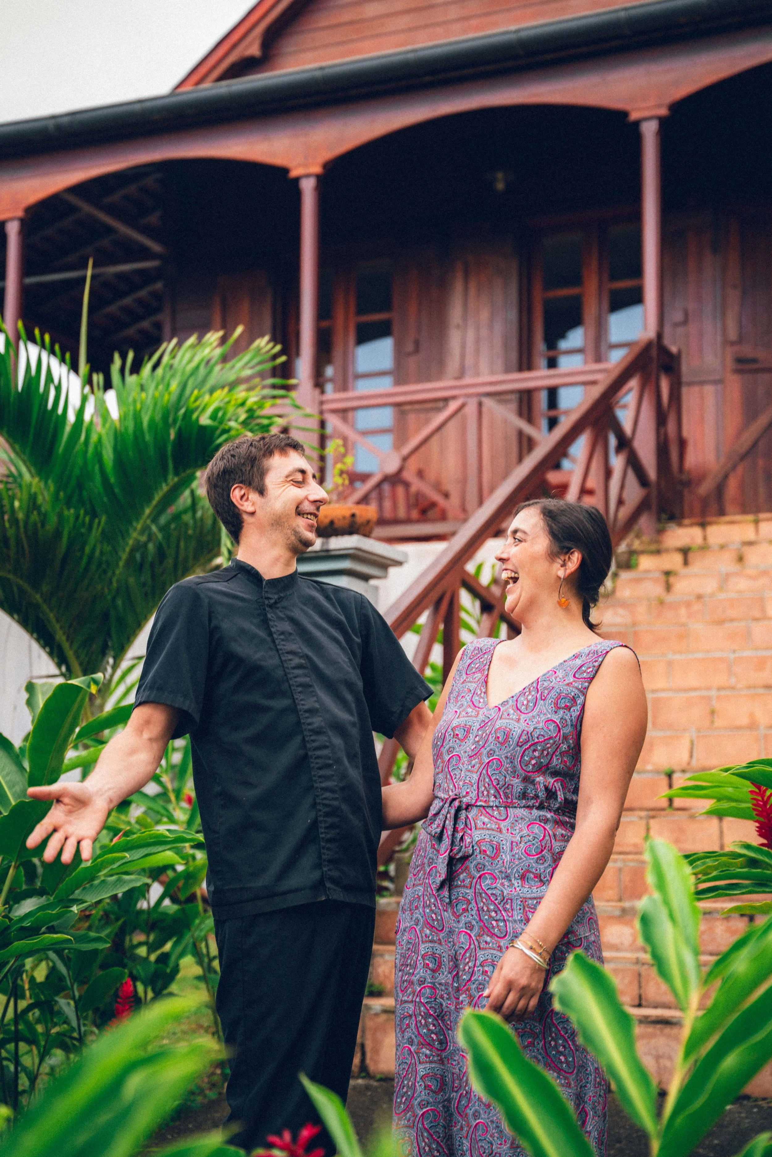 Un homme et une femme souriants, se tenant la main dans un jardin, devant une maison en bois, entourés de plantes vertes.