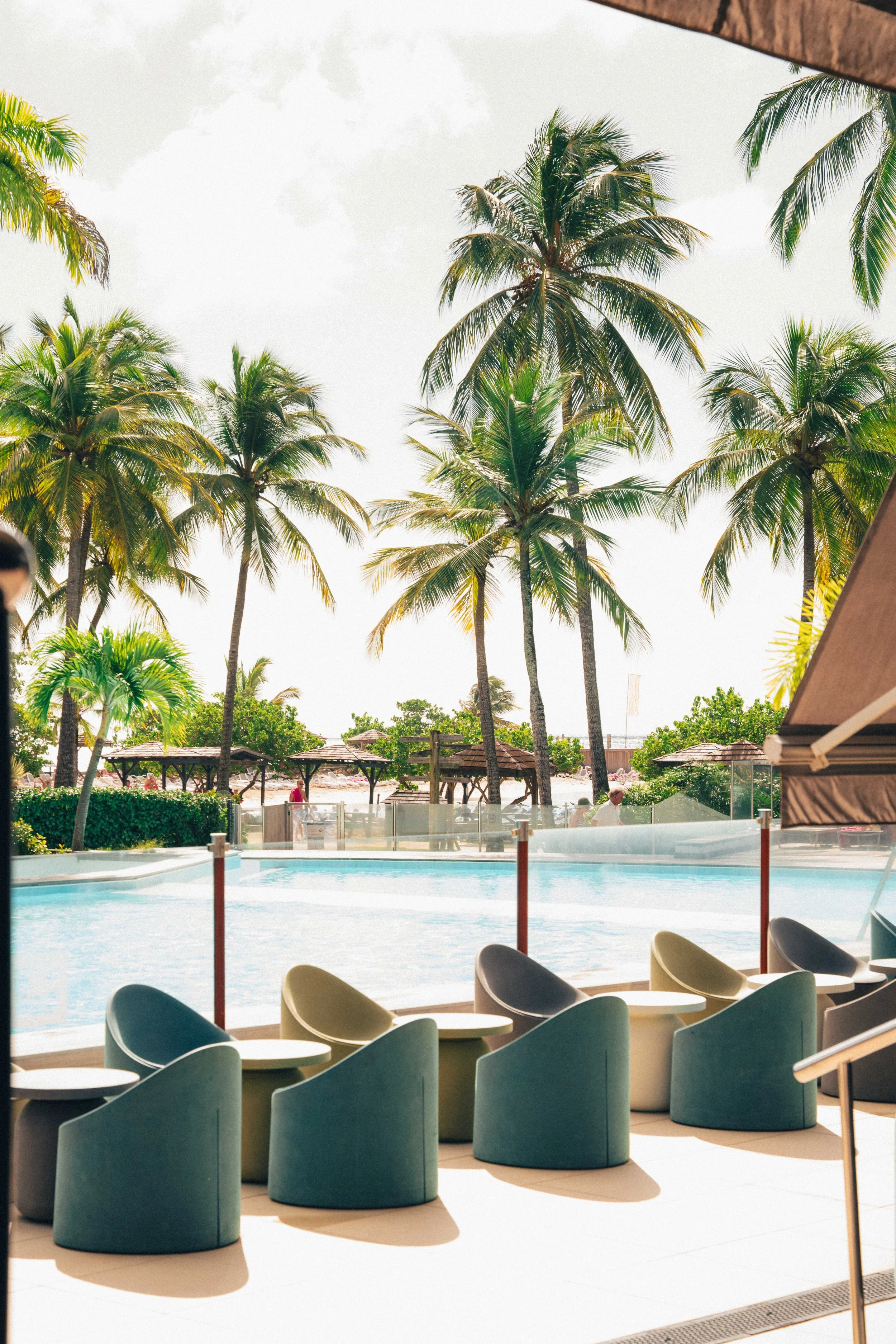 Vue depuis une salle de repos avec des chaises modernes en face d'une piscine, entourée de palmiers et de parasols sur une journée ensoleillée.