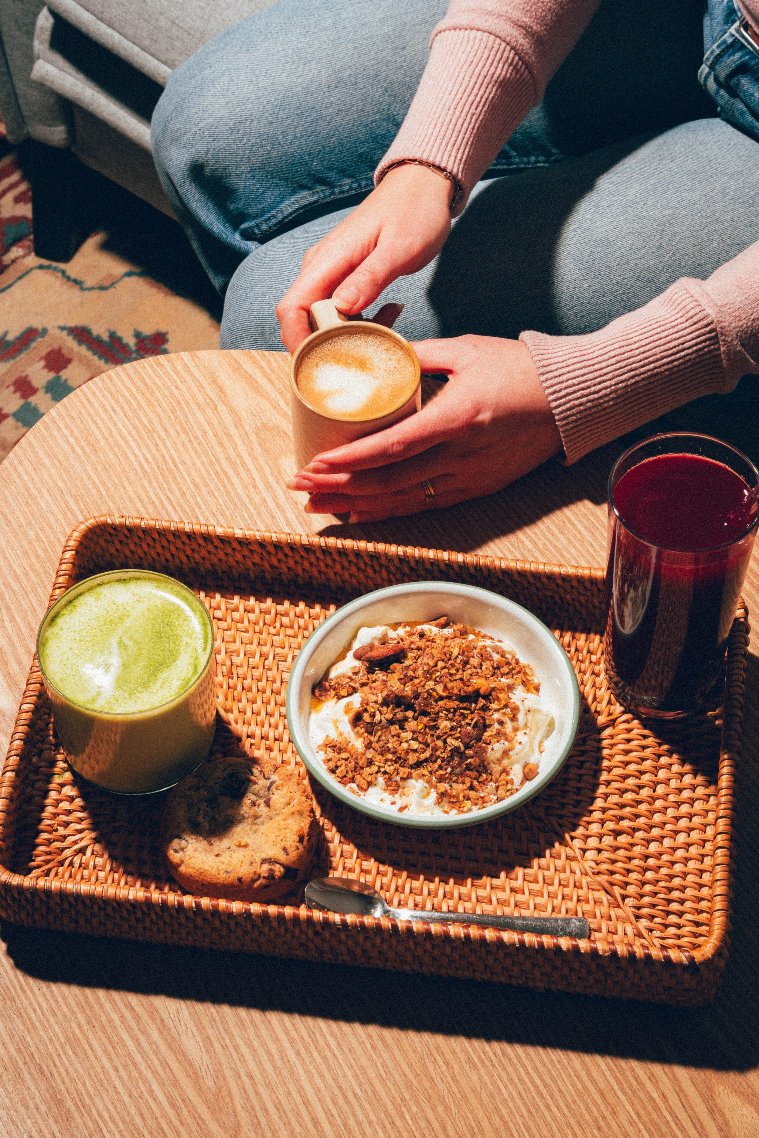 Une personne tenant une tasse de café dans un salon, avec un plateau contenant un smoothie vert, un bol de yaourt avec granola, un cookie, ainsi qu'un verre de jus rouge sur une table en rotin.