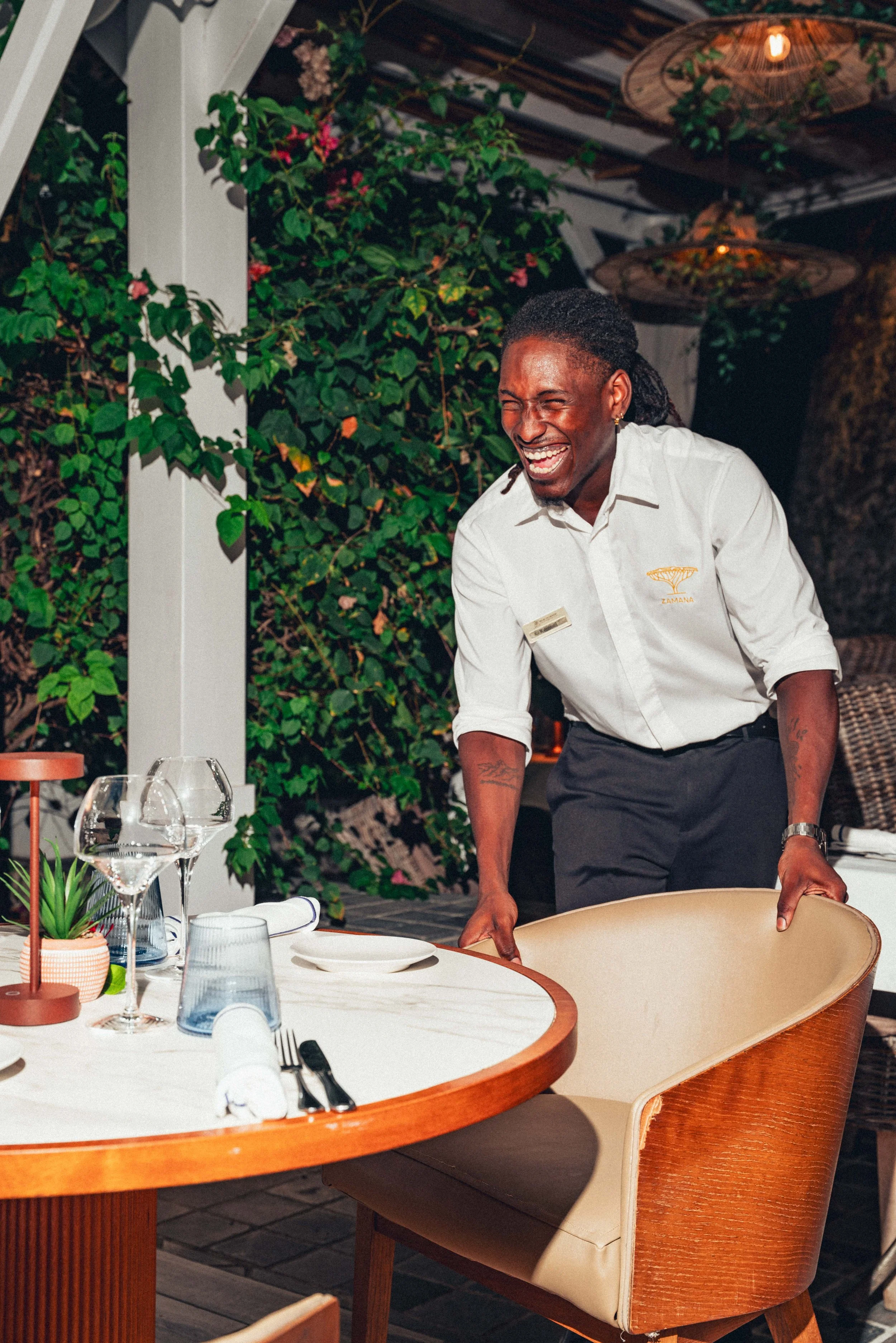 Photographie culinaire d'un serveur souriante portant une chemise blanche de serveur derrière une table de restaurant, décorée de plantes et de vaisselle, dans un cadre extérieur avec luminaire suspendu.