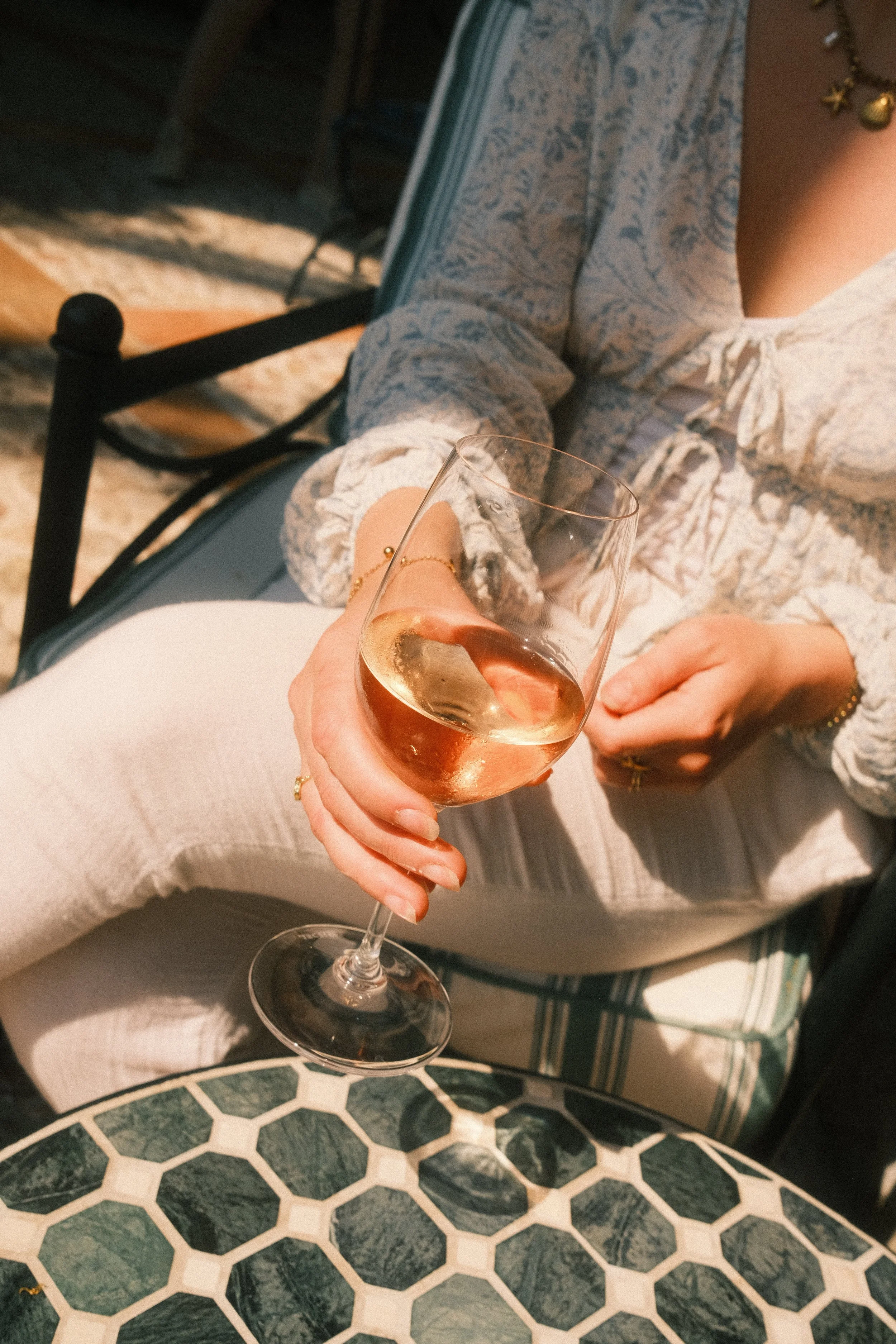 Une personne tient un verre de rosé, assise à une table en outdoor, avec un motif en carreaux de marbre blanc et vert. La personne porte un bracelet doré et une robe ou haut en tissu léger avec motif. Autre personne à côté, brun clair, portant une ch