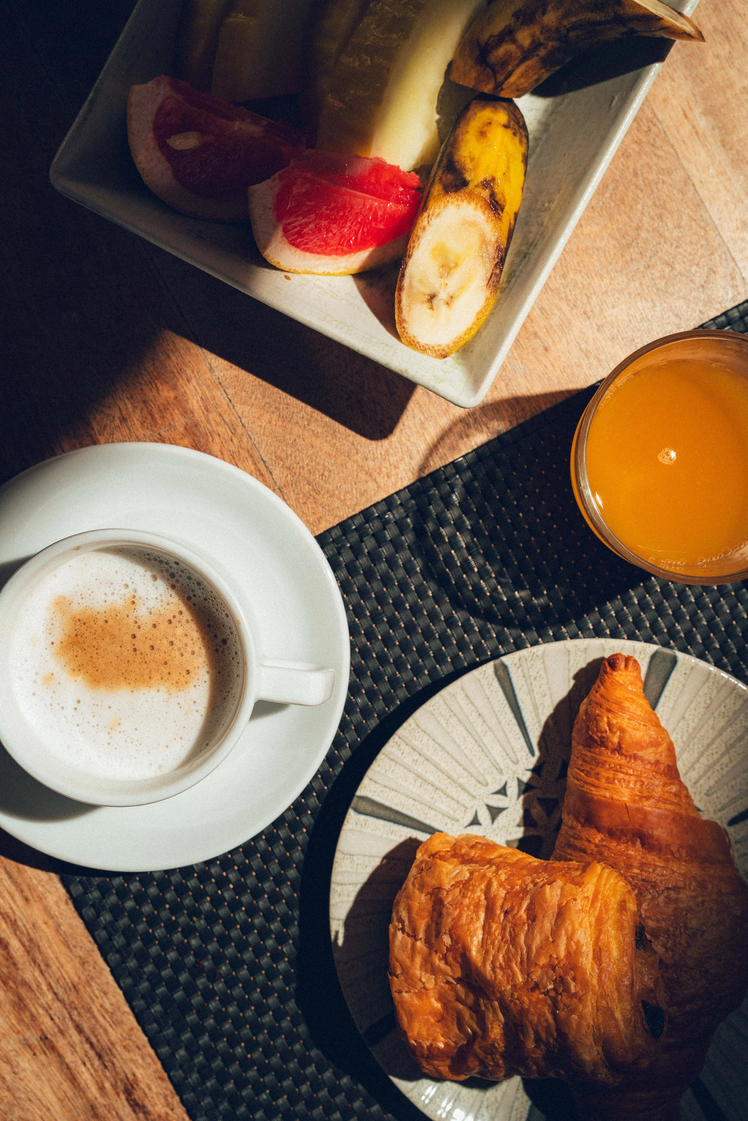 Un petit-déjeuner comprenant un croissant, deux muffins, un verre de jus d'orange, une tasse de café et un plateau avec divers fruits