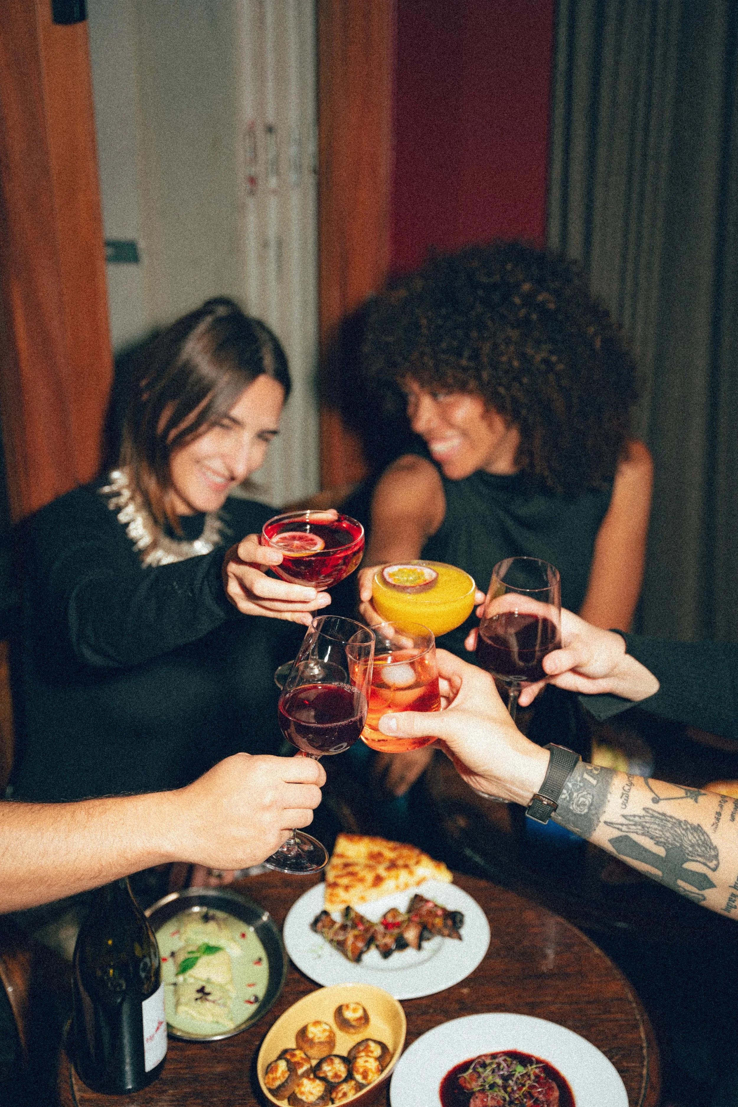 Groupe de personnes à une fête qui trinquent avec des cocktails, autour d'une table avec nourriture, y compris une pizza, salade, et des amuse-bouches.
