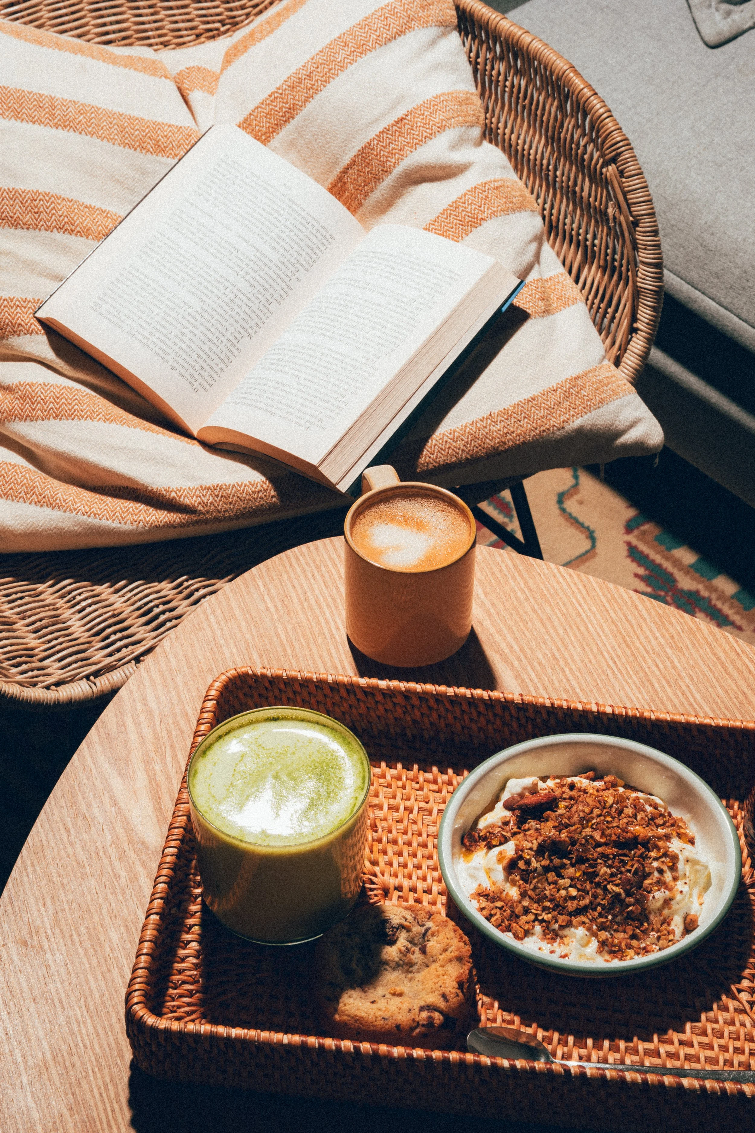 Un plateau en osier avec un bol de yaourt garni de granola, un biscuit, un verre de smoothie vert et un café dans une tasse en carton. En arrière-plan, une table ronde en bois avec un livre ouvert posé sur une couverture à rayures orange et beige, un
