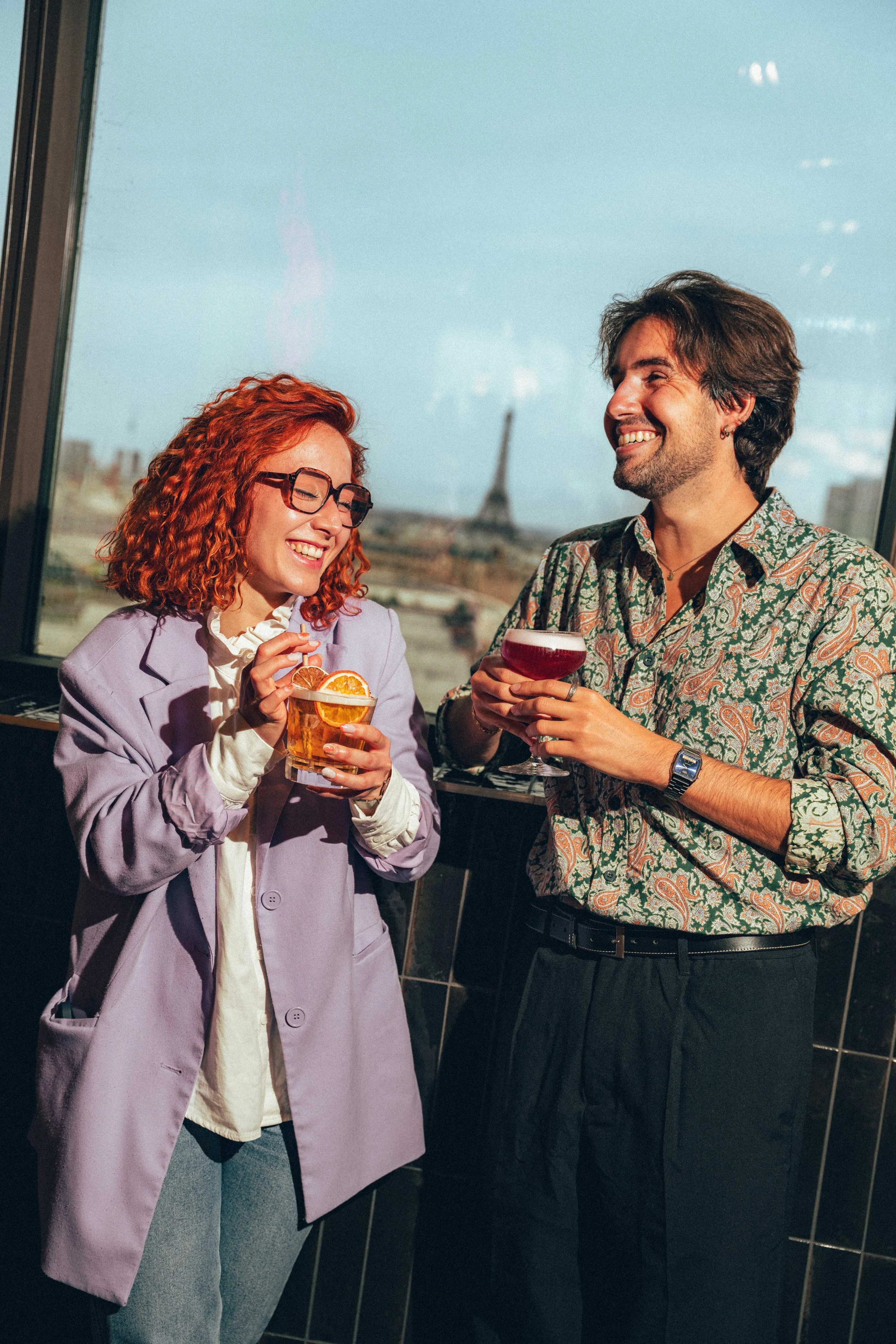 Deux personnes souriantes tenant des cocktails dans un intérieur avec vue sur la Tour Eiffel en arrière-plan.