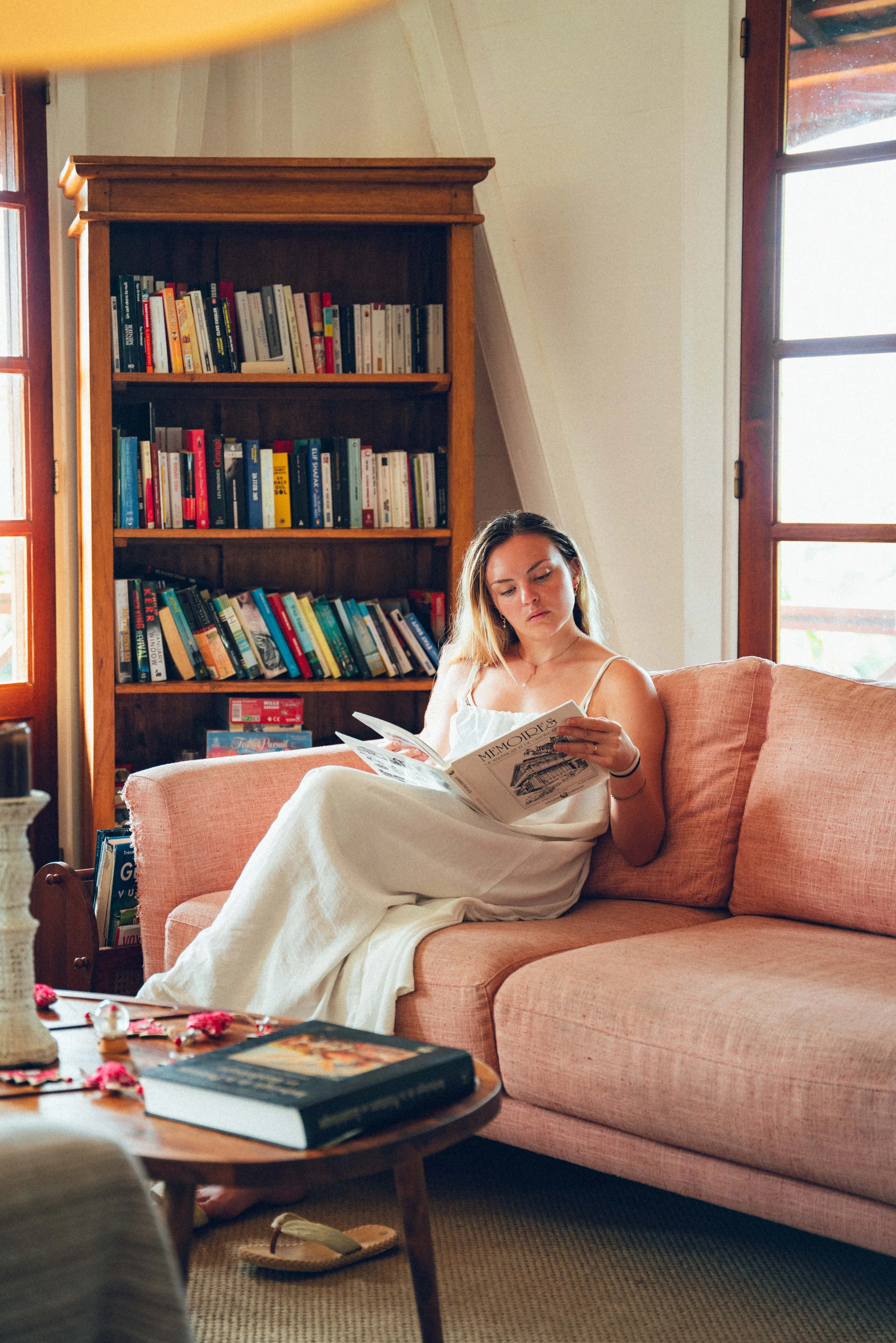 Jeune femme assise sur un canapé en tissu rose clair, lisant un magazine, dans une pièce lumineuse avec une grande bibliothèque en bois et de grandes fenêtres.